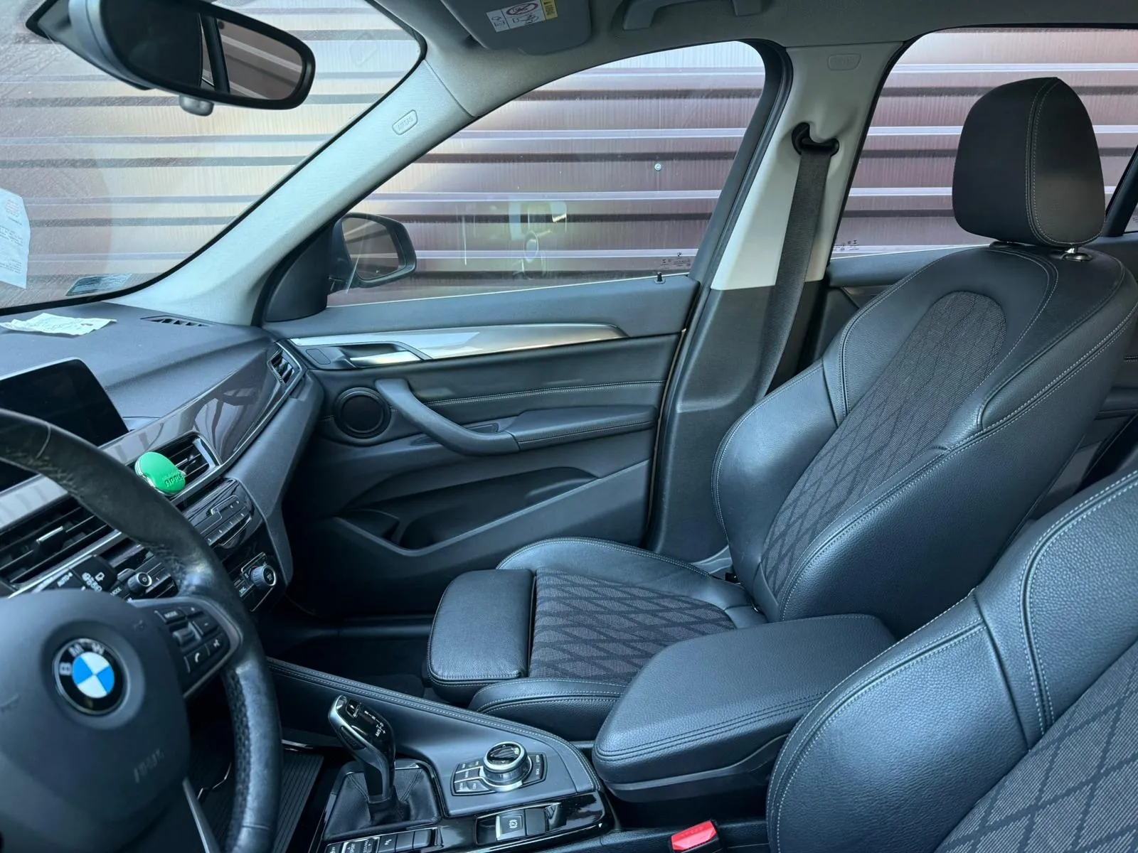 Wnętrze samochodu marki BMW z czarnymi skórzonymi fotelami w geometrycznym wzorze. Widoczna kierownica z logo BMW, deska rozdzielcza z ekranem, konsola środkowa z przyciskami i dźwignią zmiany biegów. W tle przez okna widać poziome linie budynku. Na desce rozdzielczej znajduje się zielony naklejony element. Lusterko wsteczne i pas bezpieczeństwa są w pozycji pionowej.