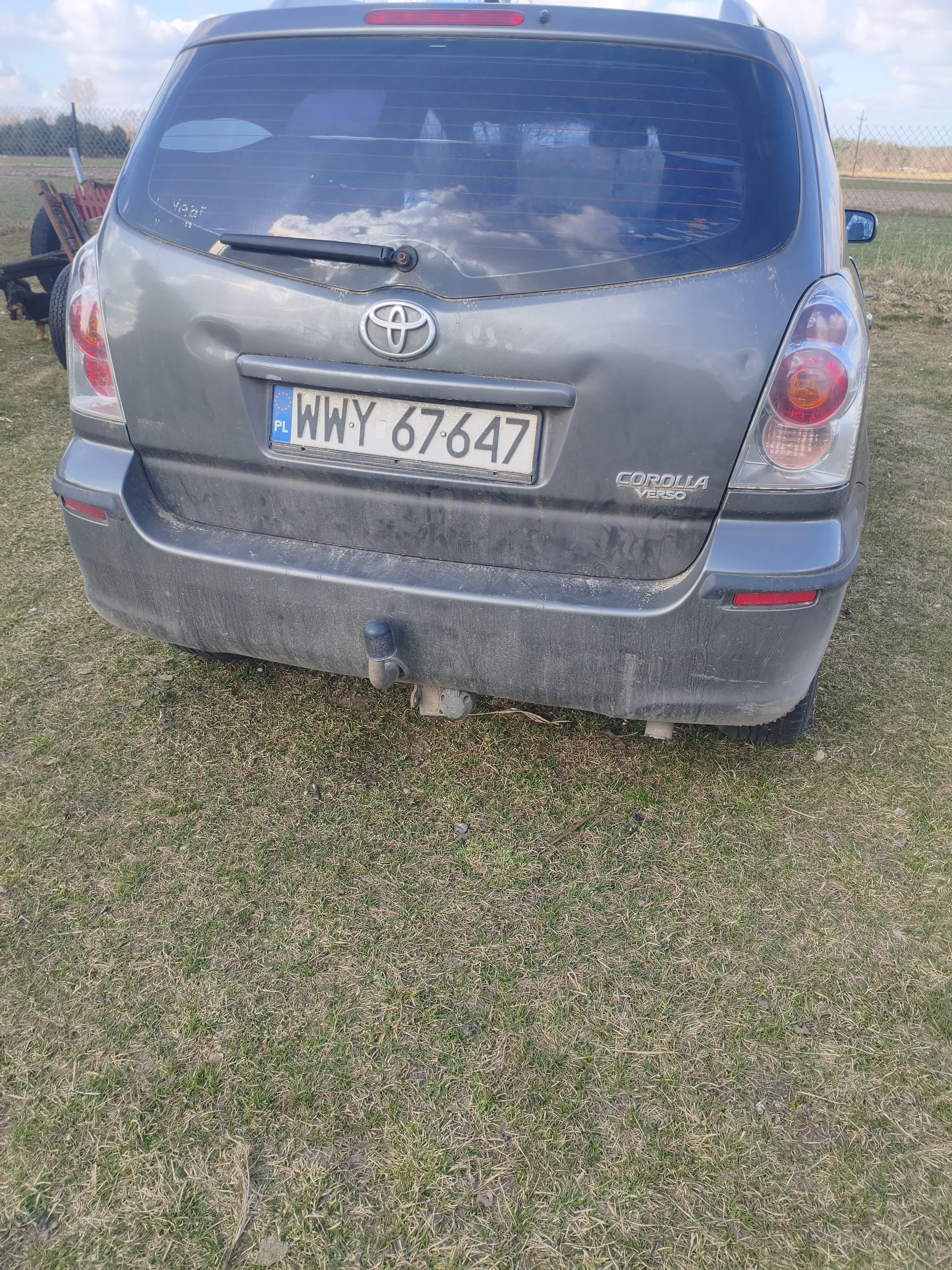 Tył szarego samochodu Toyota Corolla Verso z tablicą rejestracyjną WWY 67647 PL. Samochód jest zabrudzony, widoczny jest emblemat marki, napis "COROLLA VERSO" oraz holownik. Tło stanowi trawiaste pole, ogrodzenie i fragment maszyny rolniczej po lewej stronie. Na niebie znajdują się chmury.