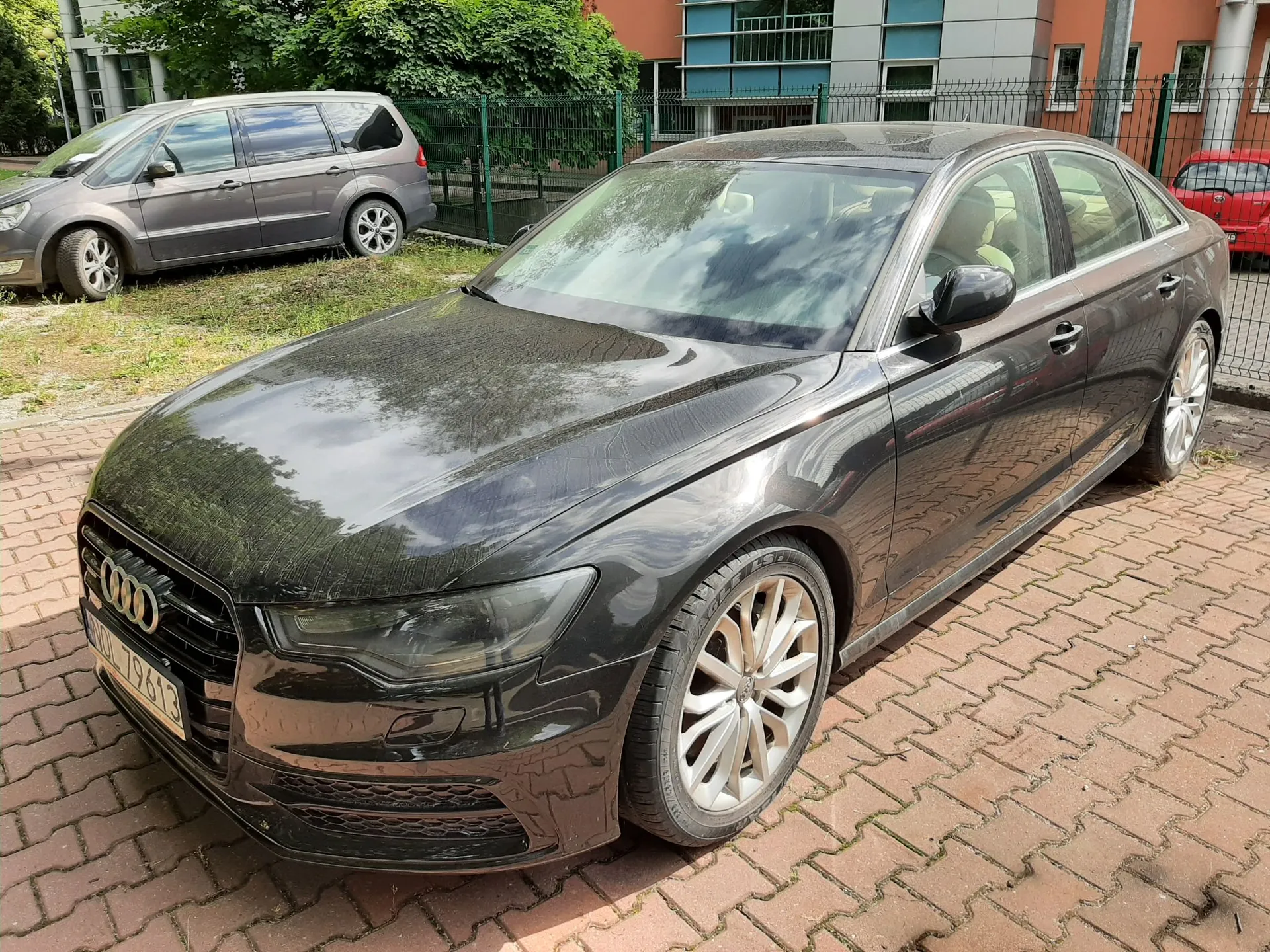 Czarny samochód marki Audi zaparkowany na kostce brukowej. Auto jest widoczne z przodu i z boku. W tle znajduje się srebrny samochód typu minivan, zielony płot oraz fragment nowoczesnego budynku z dużymi oknami i drzewami.