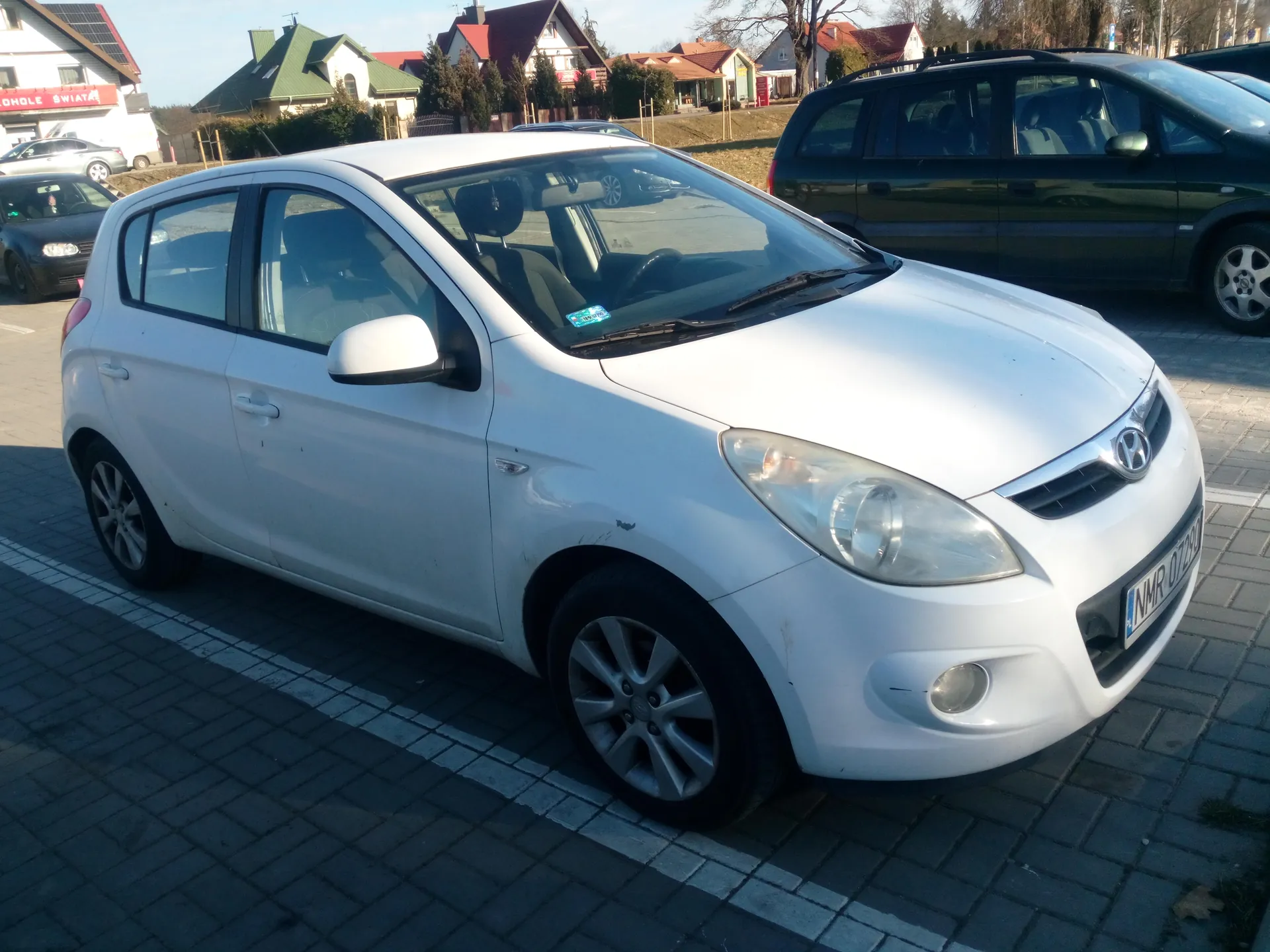 Biały samochód marki Hyundai z tablicą rejestracyjną NMR 07290 zaparkowany na brukowanym placu. W tle widoczne są inne pojazdy, domy z czerwonymi i zielonymi dachami oraz tablica z napisem OHOLE SWIAT. Na przedniej szybie samochodu znajduje się niebieski znaczek.