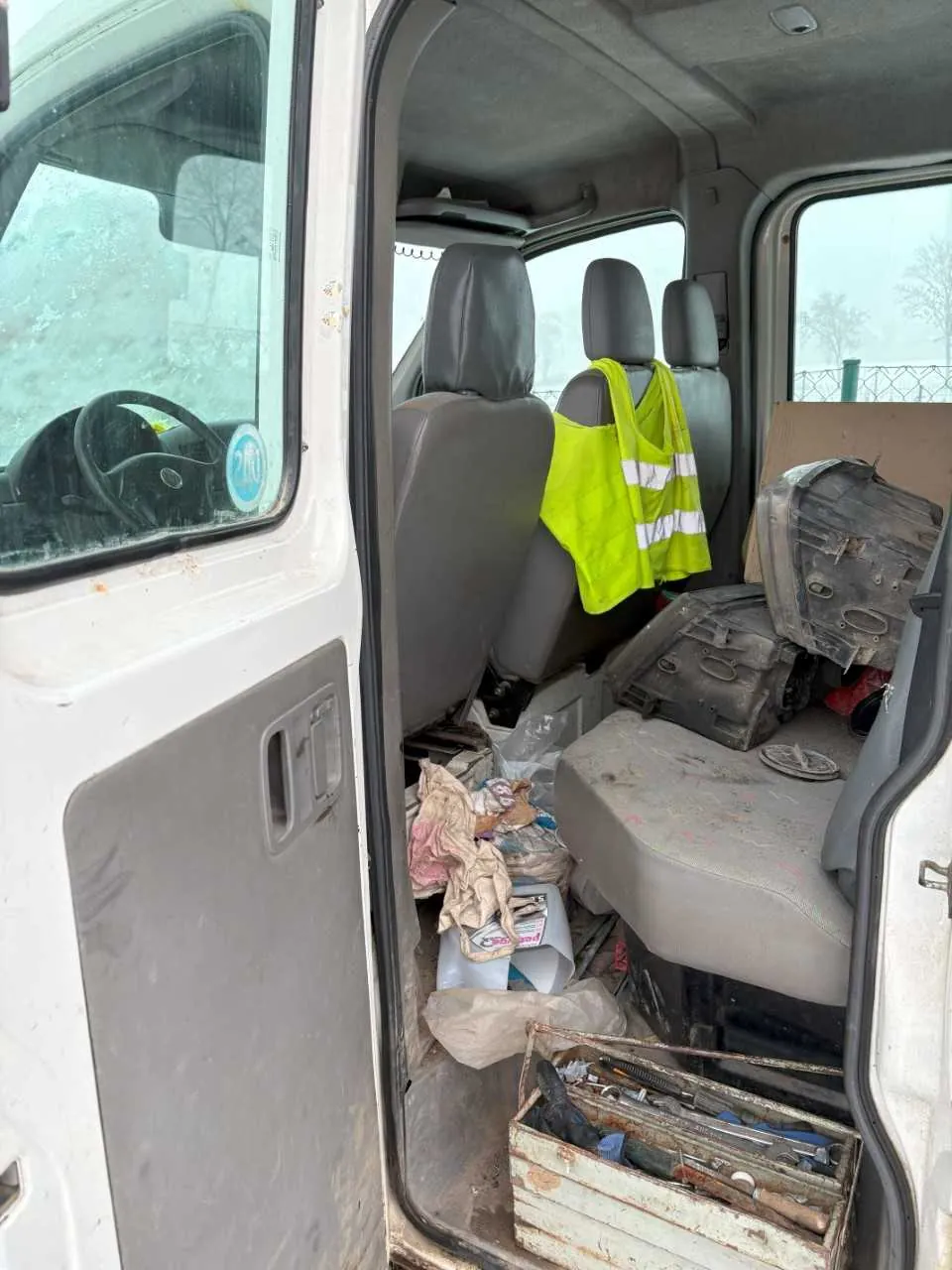 Wnętrze furgonetki z otwartymi drzwiami od strony kierowcy. Widoczne są trzy siedzenia, na środkowym wiszący jasnożółty kamizelka odblaskowa z białymi paskami. Podłoga i siedzenia są zabrudzone, rozrzucone są różne przedmioty: narzędzia w rostym zardzewiałym pudełku, worki, ubrania i inne śmieci. Przez przednią szybę widać kierownicę oraz niebieski naklejony znak. Poza pojazdem widoczny jest ogrodzenie i drzewa w mglistym otoczeniu.