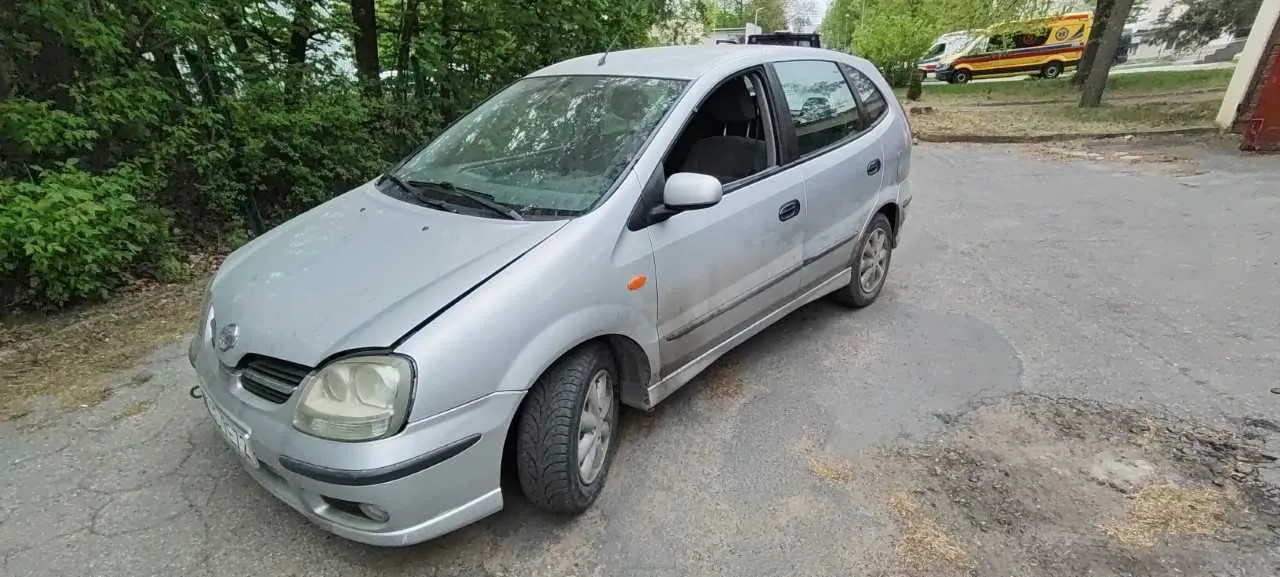Srebrny samochód osobowy typu hatchback zaparkowany na asfaltowej powierzchni. Pojazd ma widoczne ślady brudu i zużycia. W tle zielone krzewy, drzewa, fragment ogrodzenia oraz żółto-czerwony ambulans. Na pierwszym planie asfalt z plamami i suchą trawą. Samochód znajduje się w pozycji ukośnej względem krawędzi kadru.
