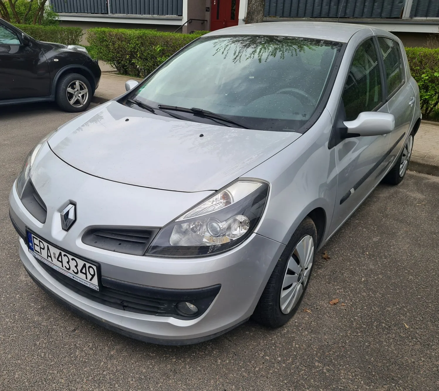 Srebrny samochód marki Renault zaparkowany na asfaltowej powierzchni. Widoczna tablica rejestracyjna z napisem EP A 43349 oraz niebieskim pasem z oznaczeniem PL po lewej stronie. W tle czarny samochód, zielone krzewy, drzewo i budynek z czerwonymi drzwiami oraz ciemnymi okiennicami. Samochód znajduje się na pierwszym planie, z przodu i boku widocznymi elementami nadwozia.