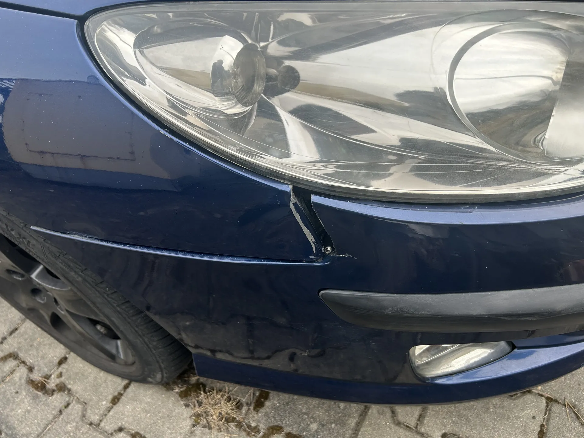 Zbliżenie przedniej części ciemnoniebieskiego samochodu. Widoczny jest reflektor, uszkodzony zarys zderzaka z widoczną pęknięciem lub zadrapaniem w okolicy połączenia z reflektorem. Część koła z oponą i felgą oraz brukowana powierzchnia z suchą trawą w szczelinach. Brak czytelnego tekstu.