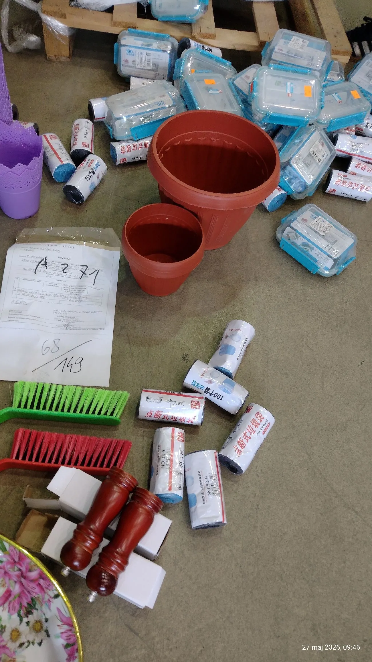 Zdjęcie przedstawiające różnorodne przedmioty rozrzucone na szarej podłodze. W górnej części widoczne są liczne przezroczyste plastikowe opakowania z niebieskimi elementami oraz białe rurki z napisami w języku chińskim. W centrum znajdują się dwie brązowe, plastikowe doniczki o różnych rozmiarach. Po lewej stronie leży biała kartka z odręcznymi zapisami, w tym numerem 'A 2771' oraz '68/149'. Poniżej znajdują się dwie szczotki z gęstym włosiem – jedna zielona i jedna czerwona. W dolnej części obrazu widoczne są dwa drewniane młynki do przypraw, białe kartonowe pudełka oraz fragment kolorowego talerza w kwiaty. W prawym dolnym rogu znajduje się data '27 maj 2026, 09:46'.