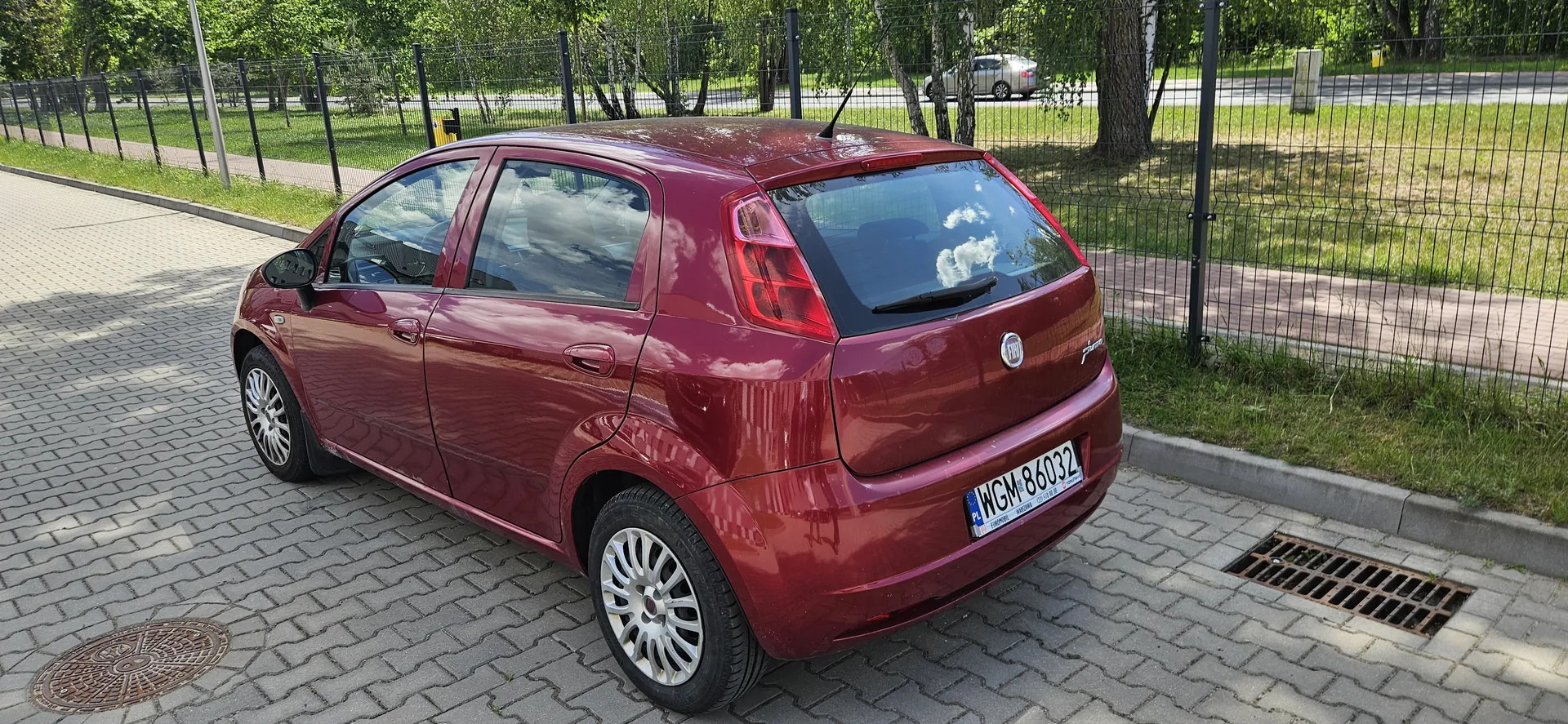 Czerwony samochód Fiat Grande Punto zaparkowany na brukowanej powierzchni. Widoczny tył pojazdu z tablicą rejestracyjną WGM 86032. W tle czarny metalowy płot, zielone drzewa, trawa oraz droga z innym samochodem w oddali. Na asfalcie znajdują się kanałowe pokrywy i żłobek odpływowy.