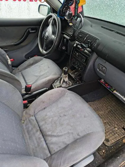 Wnętrze samochodu z przodu, widoczne zniszczone szare siedzenia, kierownica po lewej stronie, centralna konsoła z przyciskami i radio, dźwignia zmiany biegów oraz brudne dywany podłogowe. Na lusterku wstecznym wiszą ozdoby. Szyby są zabrudzone, a całość wnętrza wygląda na zużyte i nieczyste.