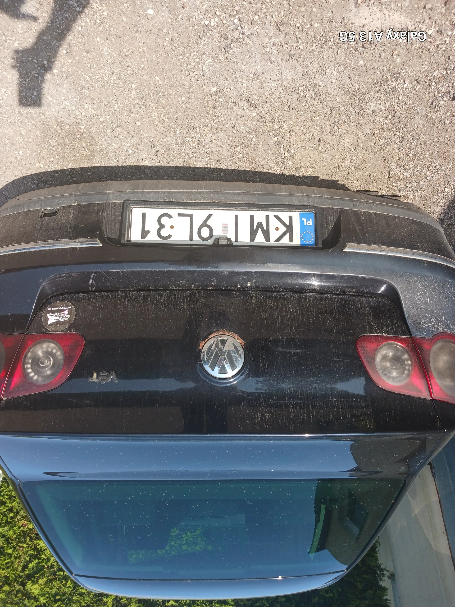 Tył czarnego samochodu marki Volkswagen z pionowo ustawioną tablicą rejestracyjną KMI 9L31. Widoczne są czerwone światła tylne, logo Volkswagena, naklejka na bagażniku oraz asfaltowa powierzchnia. Po prawej stronie obrazu znajduje się pionowy napis 'Galaxy A13 5G'.