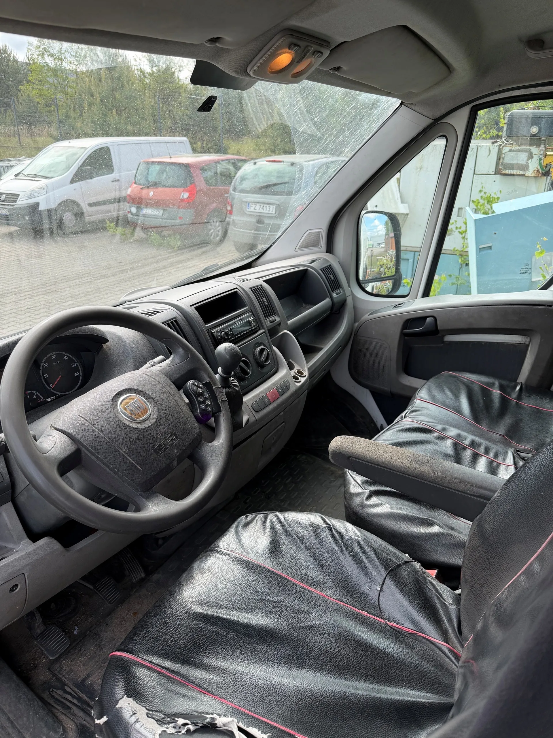 Wnętrze samochodu marki Fiat z widocznym kierownicą, deska rozdzielcza z radioodtwarzaczem i przyciskami. Czarne fotele z czerwonym przeszyciem, fotel kierowcy uszkodzony (rozerwana tapicerka). Przez przednią szybę widoczne zaparkowane pojazdy: biały van, czerwony samochód osobowy i szary samochód z tablicą rejestracyjną FZ 7413. Tło z drzewami i ogrodzeniem. Wnętrze pojazdu w kolorze szarym, podłoga zabezpieczona czarną matą.
