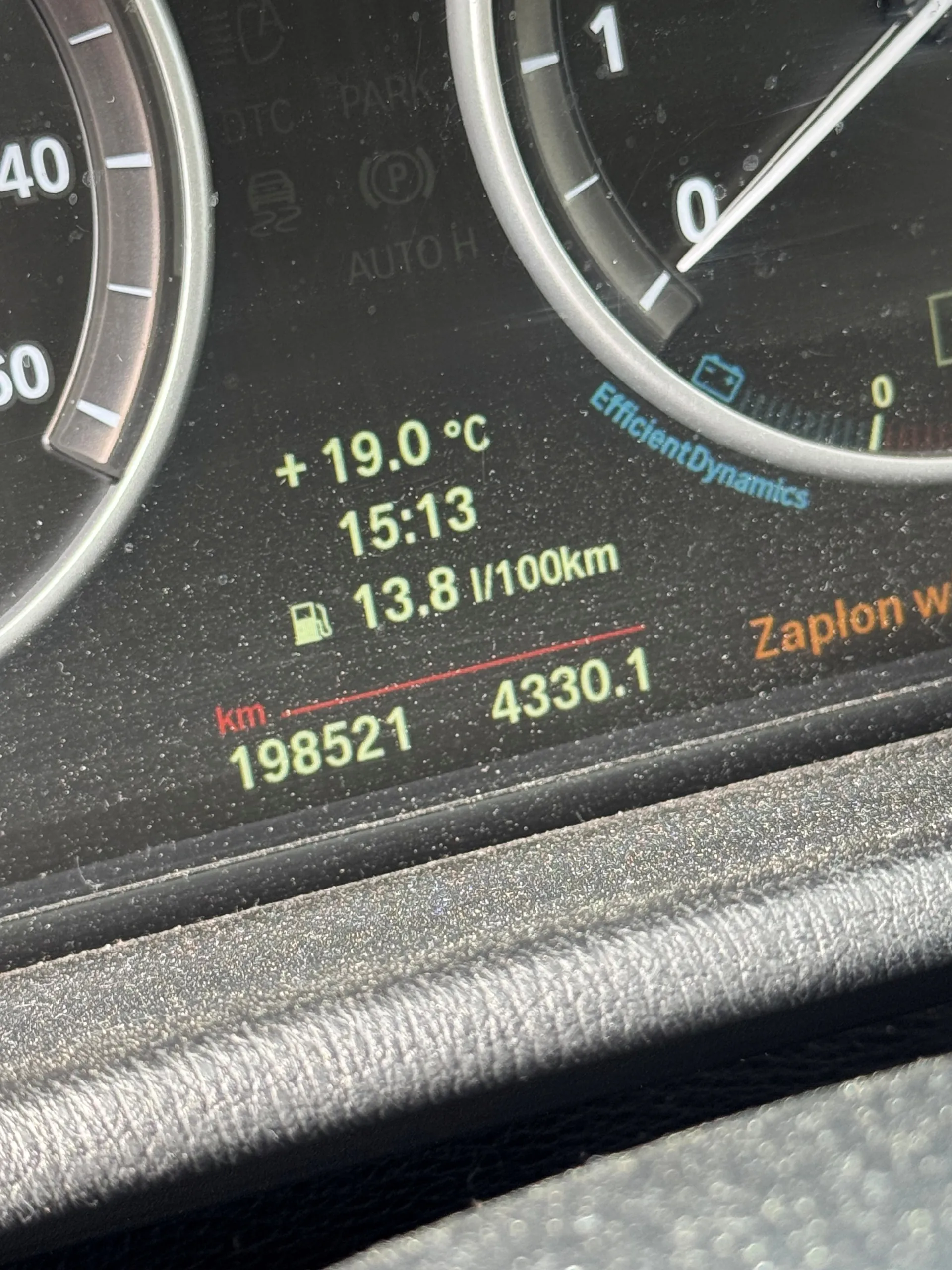 Zbliżenie na deski rozdzielcze samochodu z dwoma wskaźnikami prędkości po bokach. W centralnej części wyświetlacza cyfrowego widoczne są informacje: temperatura +19.0 °C, godzina 15:13, zużycie paliwa 13.8 l/100km, przebieg 198521 km oraz wartość 4330.1. Obok znajduje się napis 'EfficientDynamics' oraz częściowo widoczny napis 'Zaplon w'. Na wyświetlaczu widoczne są również symbole: DTC, PARK, (P), AUTO H oraz ikona paliwa.