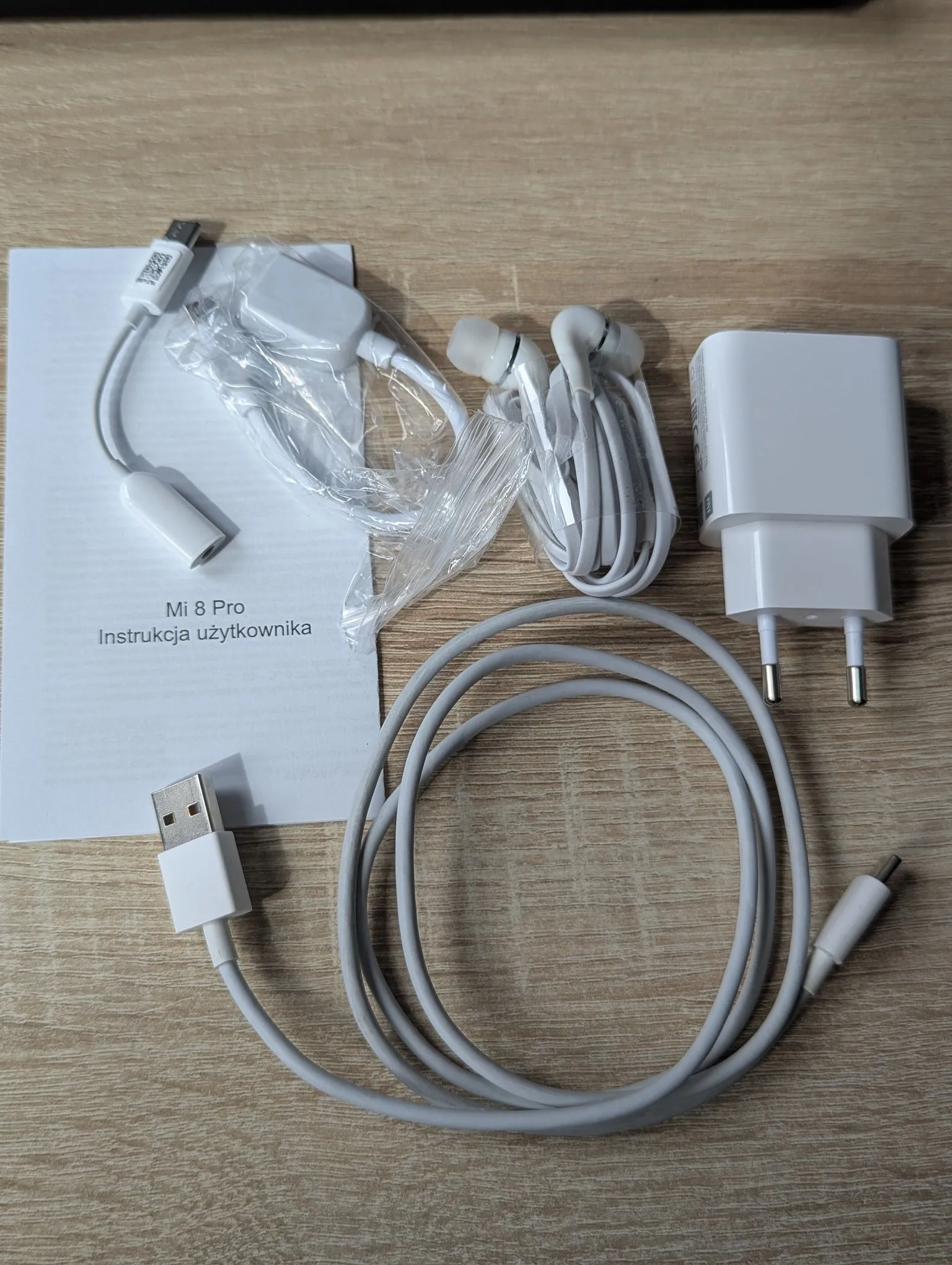 Zestaw akcesoriów elektronicznych ułożonych na drewnianym blacie. W skład zestawu wchodzi: biała instrukcja użytkownika z napisem „Mi 8 Pro Instrukcja użytkownika”, biała ładowarka sieciowa, białe słuchawki douszne z kablem, biały kabel USB do ładowania oraz dodatkowy krótki biały kabel z wtyczką USB.