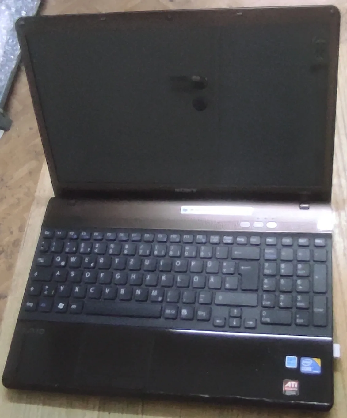 Otwarty laptop marki Sony w kolorze czarnym, ustawiony pionowo na jasnej powierzchni. Widoczny jest ekran, klawiatura QWERTY oraz gładzik. W prawym dolnym rogu obudowy znajdują się naklejki z logo Intel Core oraz ATI.