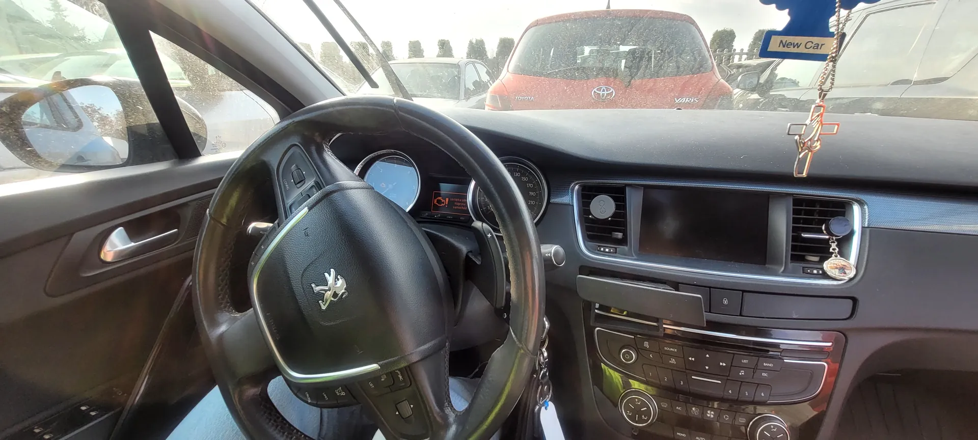 Wnętrze samochodu widziane z miejsca kierowcy. Kierownica z logo Peugeot, tablica rozdzielcza z licznikami i ostrzeżeniem na wyświetlaczu. W centralnej konsoli znajduje się radio, klimatyzacja oraz przyciski sterujące. Na lusterku wstecznym wiszą tabliczka z napisem 'New Car' oraz ozdoba w kształcie krzyża. Przez przednią szybę widoczny czerwony samochód Toyota Yaris oraz inne pojazdy na tle drzew i nieba.