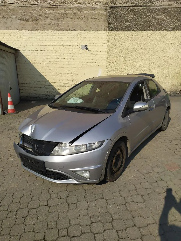 Srebrny samochód osobowy marki Honda Civic z widocznymi uszkodzeniami przodu. Maska jest wgnieciona i lekko uchylona, a przedni zderzak jest pęknięty i częściowo oderwany po prawej stronie (patrząc od przodu). Auto stoi na kostce brukowej przed jasną ścianą budynku. W tle po lewej stronie widoczny jest pomarańczowo-biały pachołek drogowy.