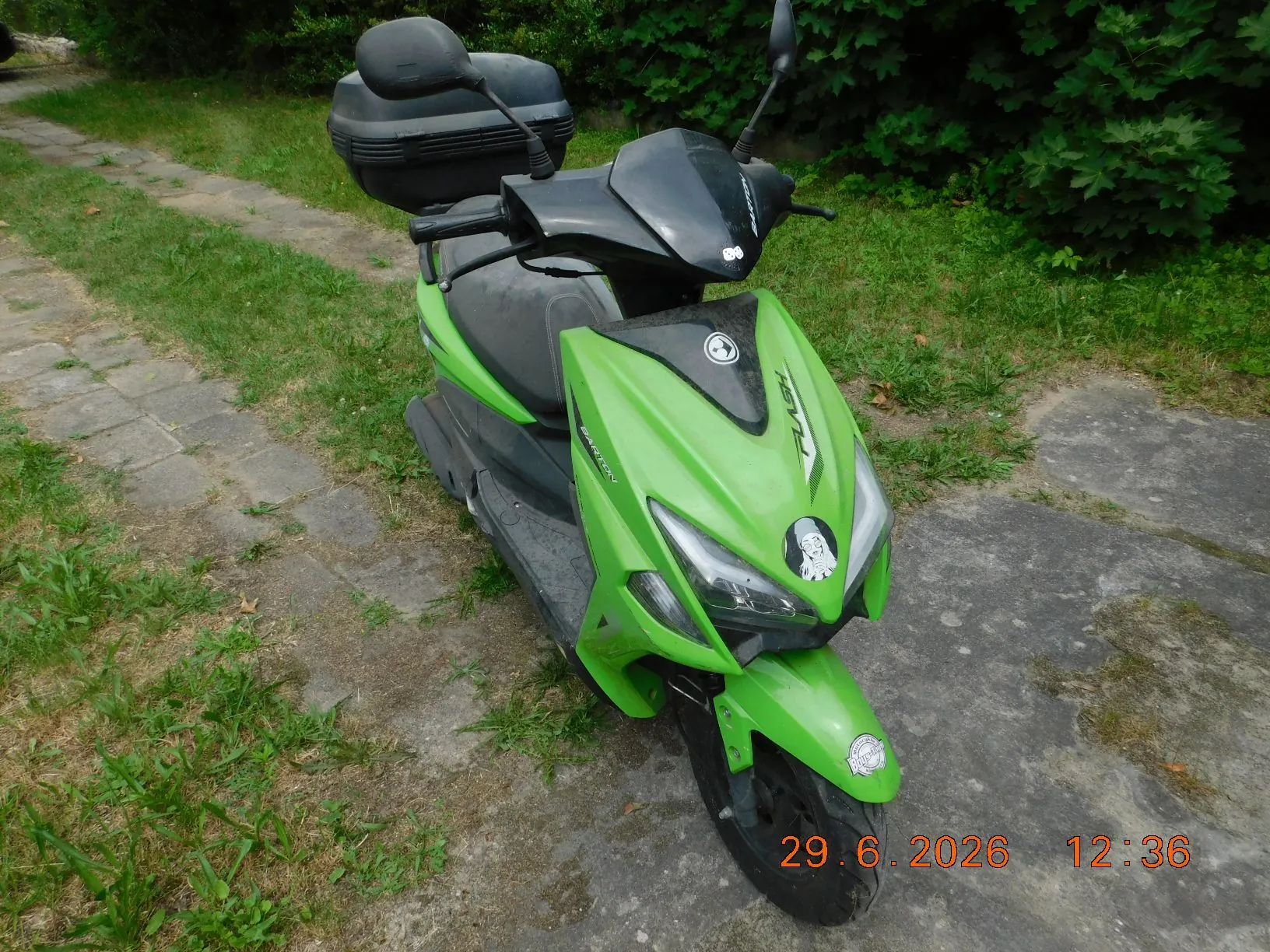 Jasnozielony skuter zaparkowany na betonowej ścieżce obok trawnika i zielonych krzewów. Skuter posiada czarny kufer z tyłu, czarną kanapę oraz czarną owiewkę nad reflektorem. Na przedniej obudowie widoczne są napisy 'FLASH' oraz 'BARTON'. W prawym dolnym rogu zdjęcia znajduje się pomarańczowa data i godzina: '29.6.2026 12:36'.