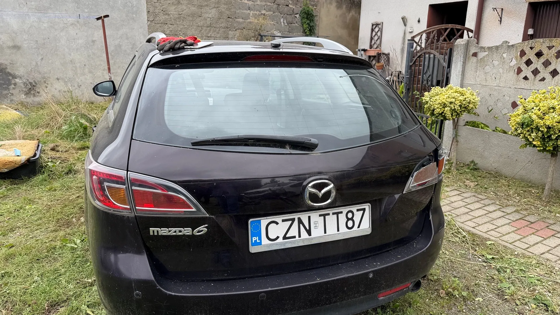 Tylny widok ciemnofioletowego samochodu marki Mazda 6. Na klapie bagażnika znajduje się logo Mazdy, napis 'mazda 6' oraz polska tablica rejestracyjna o numerze 'CZN TT87'. Na dachu samochodu leży czerwona szmatka. Auto zaparkowane jest na trawiastym podjeździe obok betonowego muru i bramy.