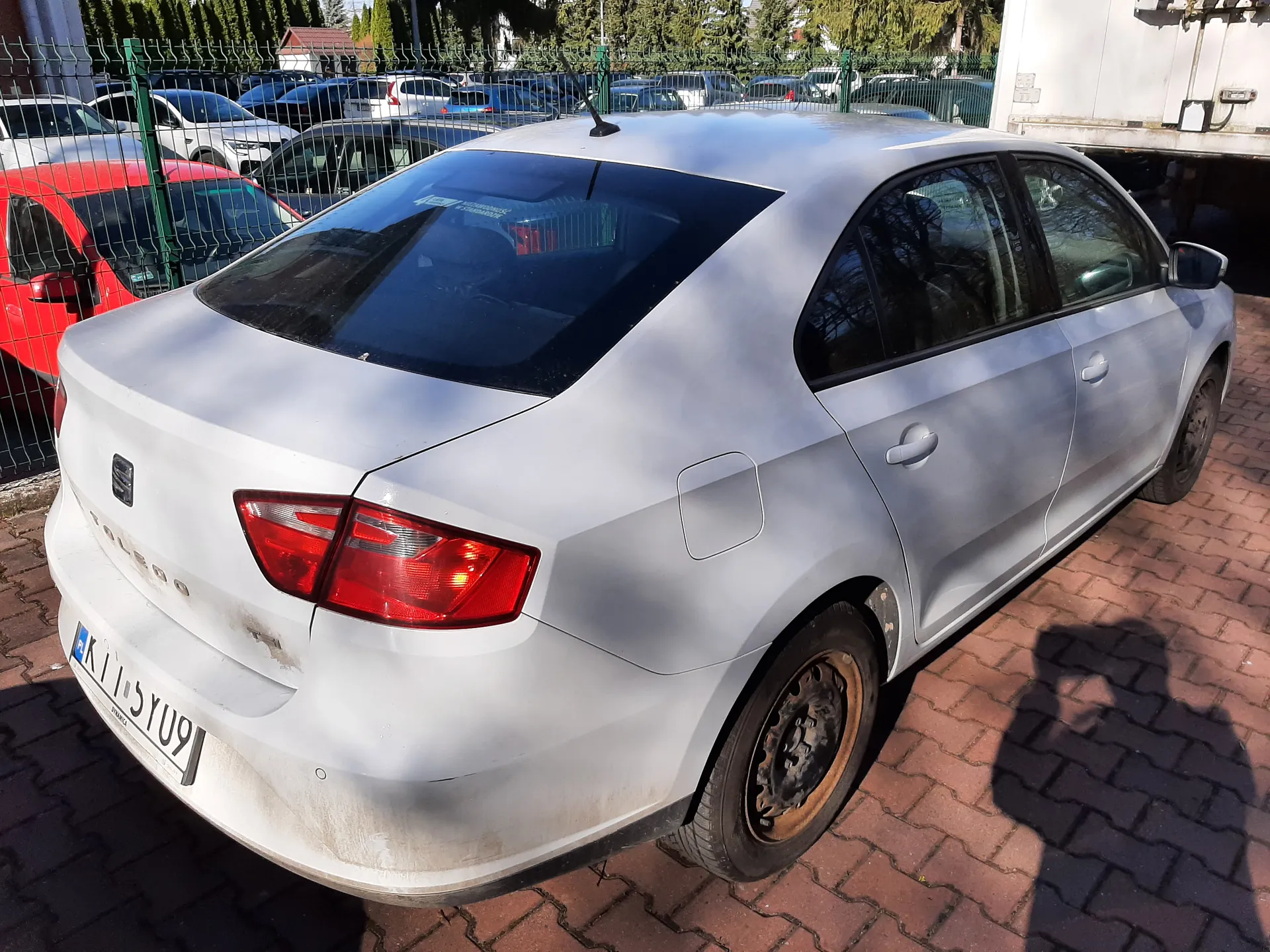 Biały samochód osobowy marki SEAT Toledo, widok z tyłu i prawej strony. Na tablicy rejestracyjnej widoczny numer KI 5YU9. Samochód stoi na ceglanym placu parkingowym, na dolnej części nadwozia i felgach widoczne zabrudzenia. W tle zielone ogrodzenie, za którym zaparkowane są inne pojazdy, w tym czerwony samochód. W oddali drzewa i budynki. Na ziemi widoczny cień osoby robiącej zdjęcie. Słoneczna pogoda, jasne oświetlenie.
