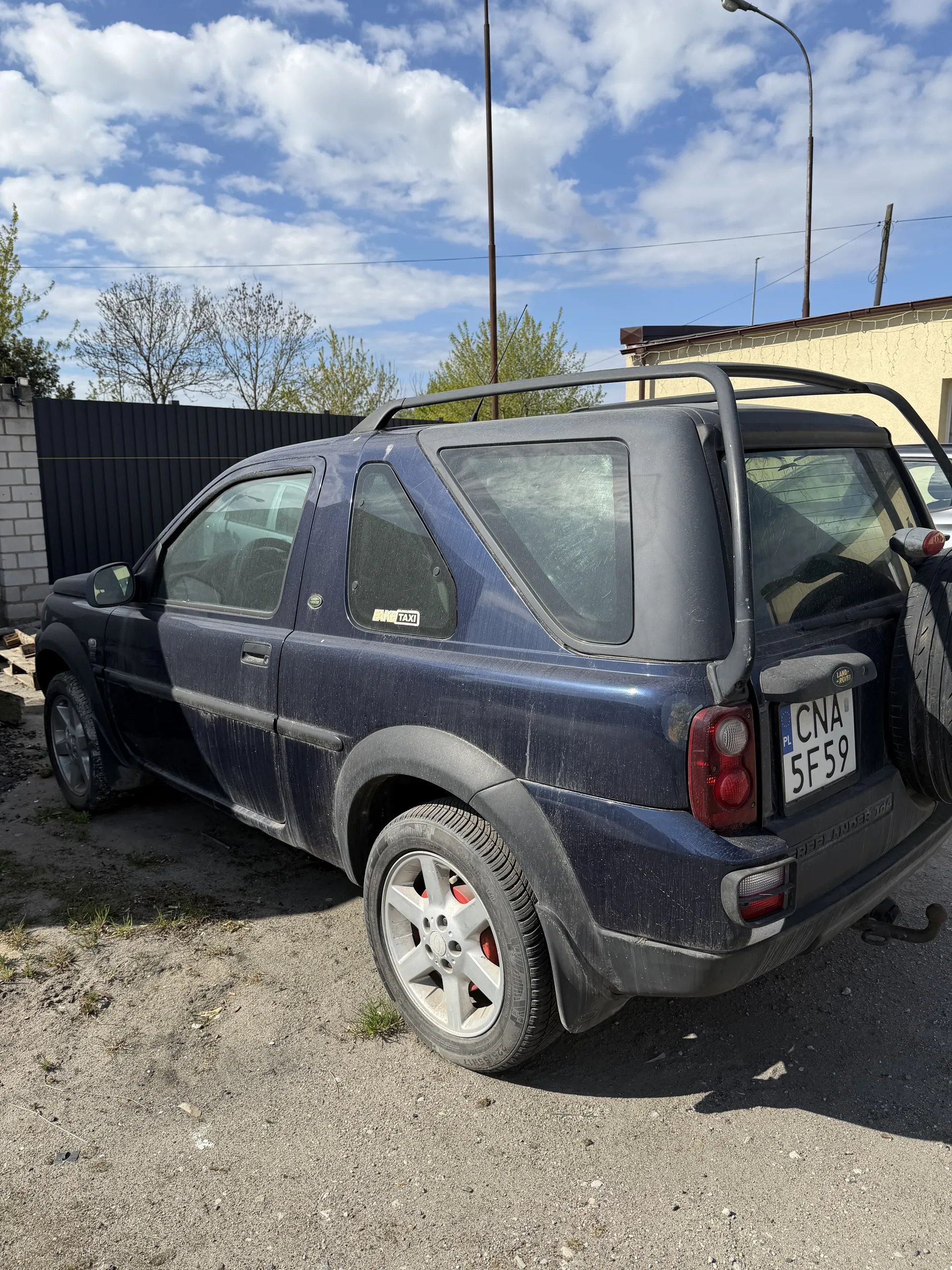 Ciemnoniebieski samochód osobowy zaparkowany na żwirowej powierzchni. Na dachu widoczny bagażnik dachowy, a na tylnym zderzaku zamontowana zapasowa opona. Tablica rejestracyjna z napisem CNA 5F59. W tle budynki, drzewa i niebo z białymi chmurami. Samochód jest lekko zabrudzony.