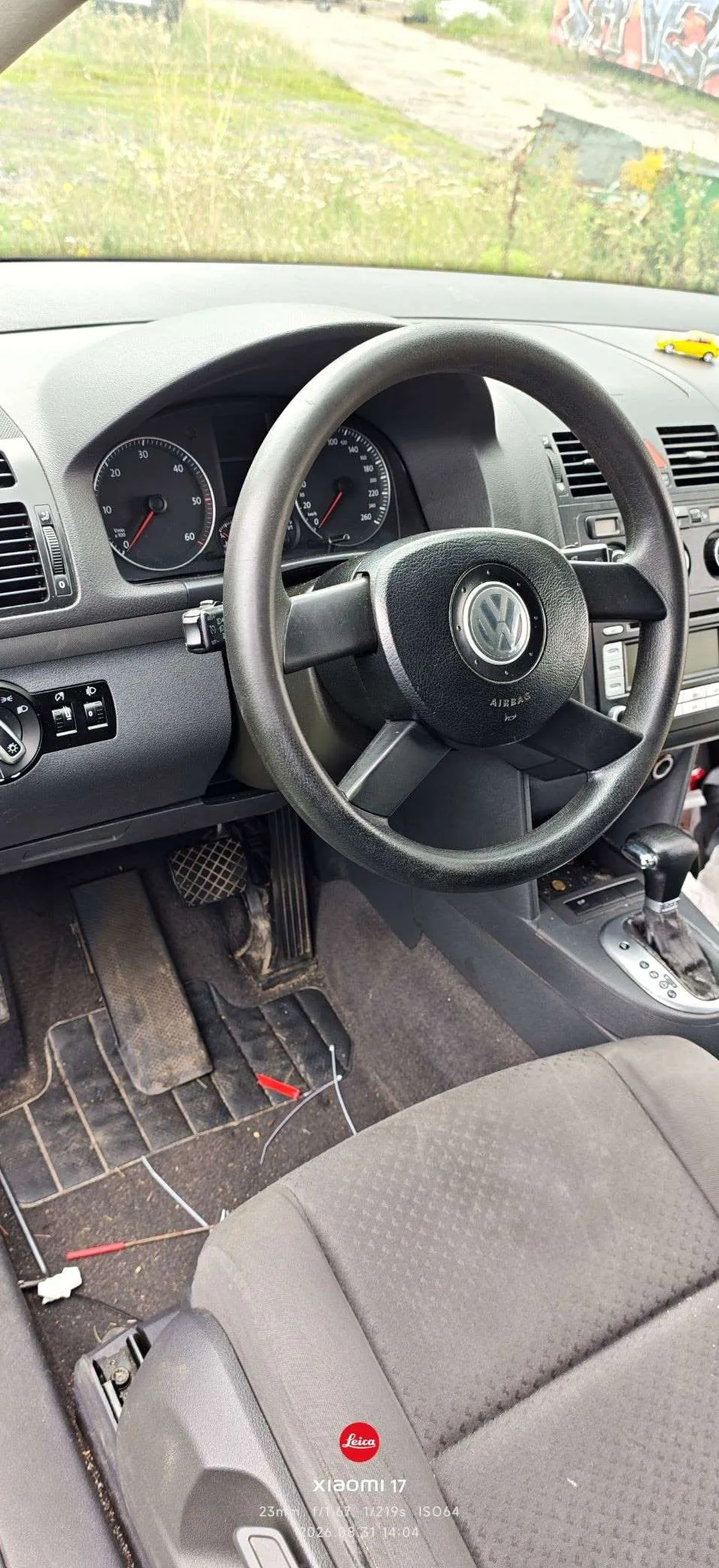 Wnętrze samochodu marki Volkswagen widziane z perspektywy kierowcy. Widoczna jest czarna kierownica z logo VW i napisem AIRBAG, deska rozdzielcza z analogowymi zegarami (prędkościomierzem i obrotomierzem) oraz konsola środkowa z lewarkiem zmiany biegów. Na podłodze po stronie kierowcy znajdują się luźne przewody elektryczne i gumowa mata. W dolnej części zdjęcia widoczny jest fragment szarego fotela oraz znak wodny z tekstem 'Leica Xiaomi 11T 23mm f/1.9 1/219s ISO64 2020-09-31 14:04'.