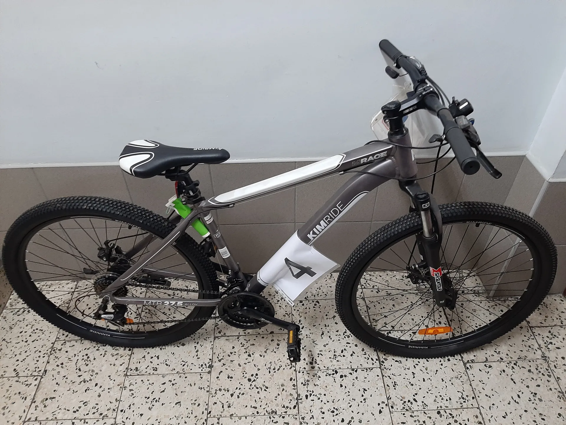 Rower górski marki KIMRIDE z ramą w kolorze szaro-białym. Na ramie widoczne napisy 'KIMRIDE', 'MIRAGE' oraz 'MTB 27.5 ALLOY'. Rower posiada czarne opony z grubym profilem, czarno-białe siedzisko, czarne kierownicę z uchwytami oraz białą tabliczkę z czarną cyfrą '4' zamocowaną na ramie. W tle szara ściana z płytkami i podłoga z kafelkami w kropki. Na przednim kole widoczny pomarańczowy odblask.