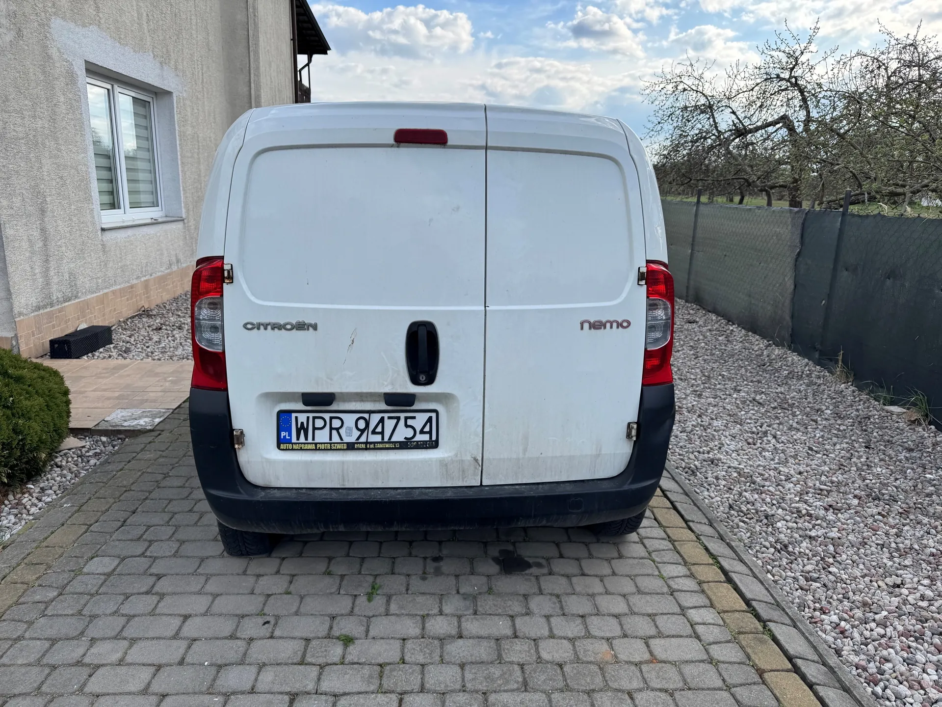 Biały samochód dostawczy Citroën Nemo z tyłu, z widoczną tablicą rejestracyjną WPR 94754 (PL) oraz napisami "CITROËN" i "nemo" na drzwiach. Pojazd stoi na brukowanej powierzchni, po lewej stronie budynek z oknem i krzewem, po prawej ogrodzenie i drzewa bez liści. Niebo częściowo zachmurzone.