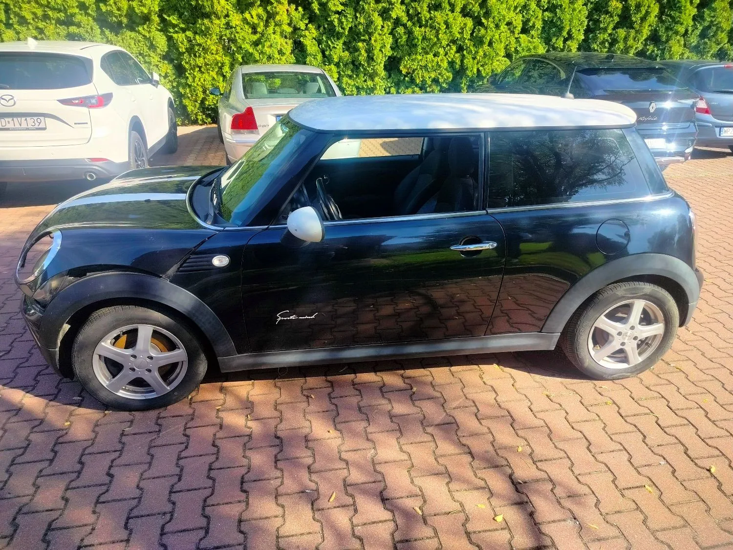Czarny samochód Mini Cooper z białym dachem i białymi lusterkami, zaparkowany bokiem na kostce brukowej. W tle widoczne są inne zaparkowane samochody: biały Mazda z tablicą rejestracyjną D 1V139, srebrny sedan oraz ciemny SUV, a za nimi gęsty, zielony żywopłot.