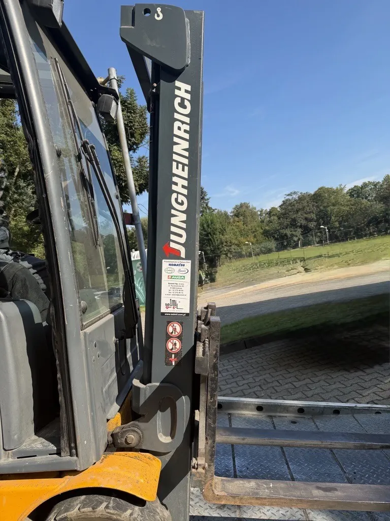 Zbliżenie na szary maszt wózka widłowego marki Jungheinrich. Na masztu widnieje pionowy biały napis 'JUNGHEINRICH' oraz naklejka z instrukcjami bezpieczeństwa i logo AUSA. W tle widoczny jest fragment kabiny operatora, zielony trawnik oraz drzewa pod jasnoniebieskim niebem.