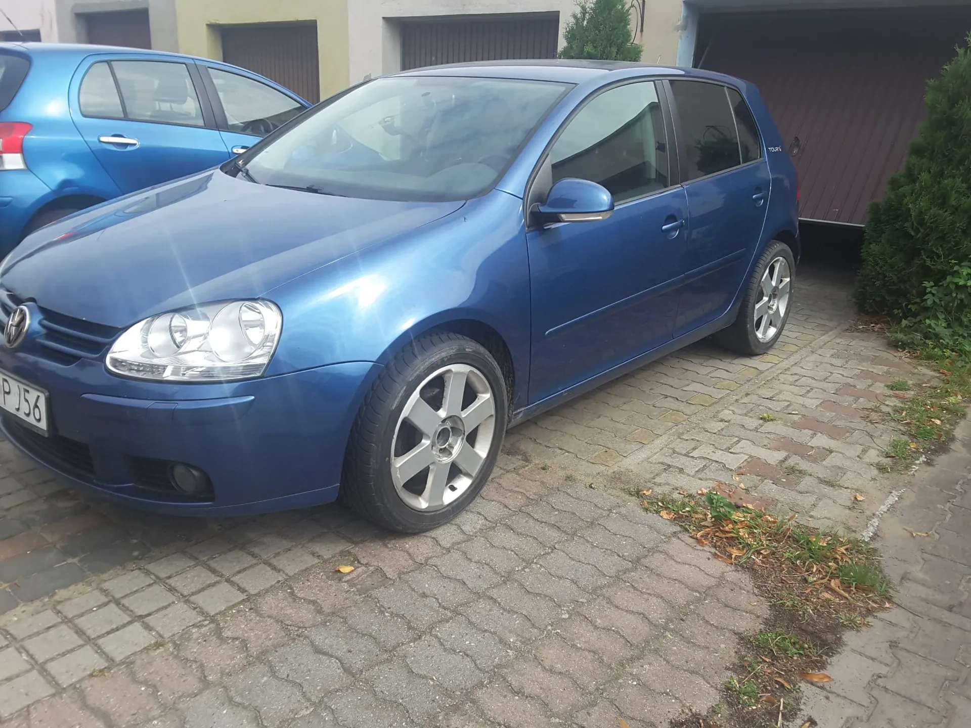 Niebieski samochód osobowy marki Volkswagen Golf zaparkowany na kostce brukowej. Auto jest widoczne z przodu i z boku. W tle widać fragment innego niebieskiego samochodu oraz budynek z garażami i zielony krzew.