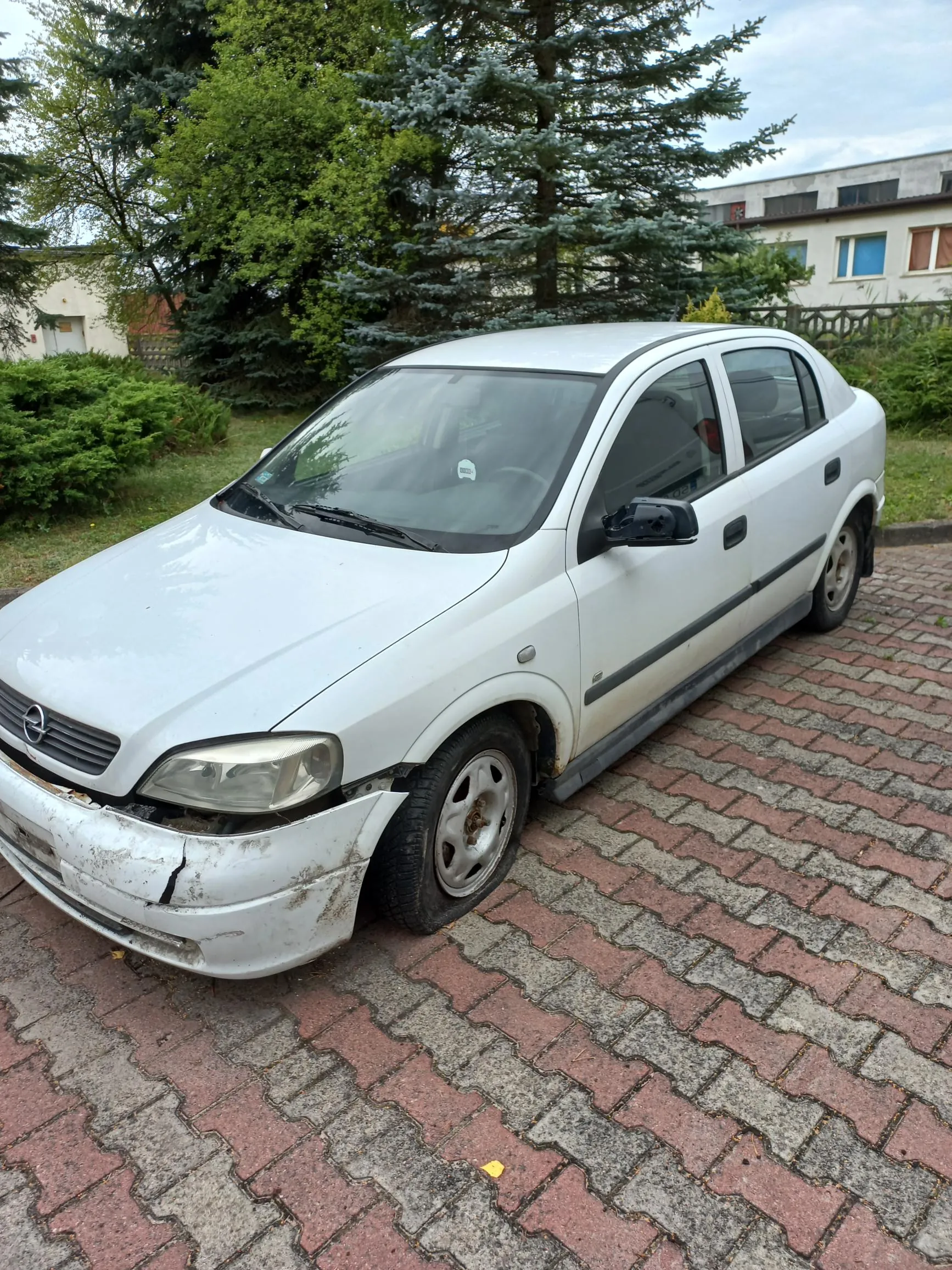 Biały samochód osobowy marki Opel zaparkowany na kostce brukowej. Pojazd ma widoczne uszkodzenia przedniego zderzaka po prawej stronie (patrząc od przodu), gdzie plastik jest pęknięty i odkształcony. W tle znajdują się zielone drzewa i fragment jasnego budynku mieszkalnego.