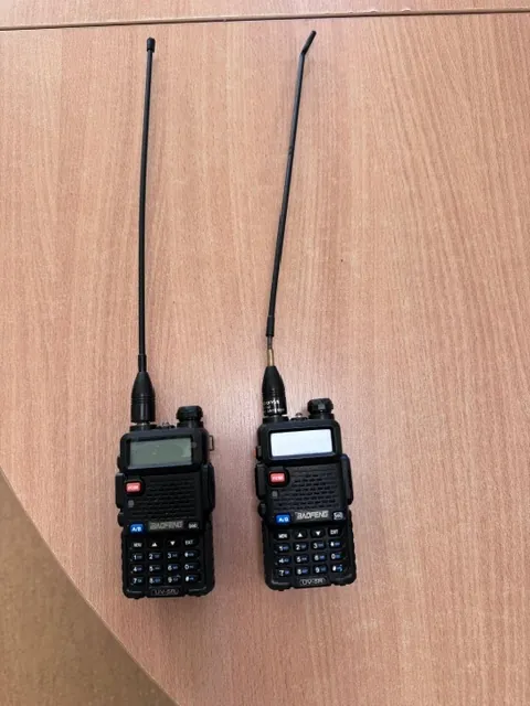 Dwa czarne radia krótkofalowe marki Baofeng model UV-5R leżące obok siebie na drewnianym blacie. Każde radio posiada długą, czarną antenę, ekran LCD oraz klawiaturę numeryczną z przyciskami. Na obudowach widoczne są napisy 'BAOFENG' oraz 'UV-5R'.