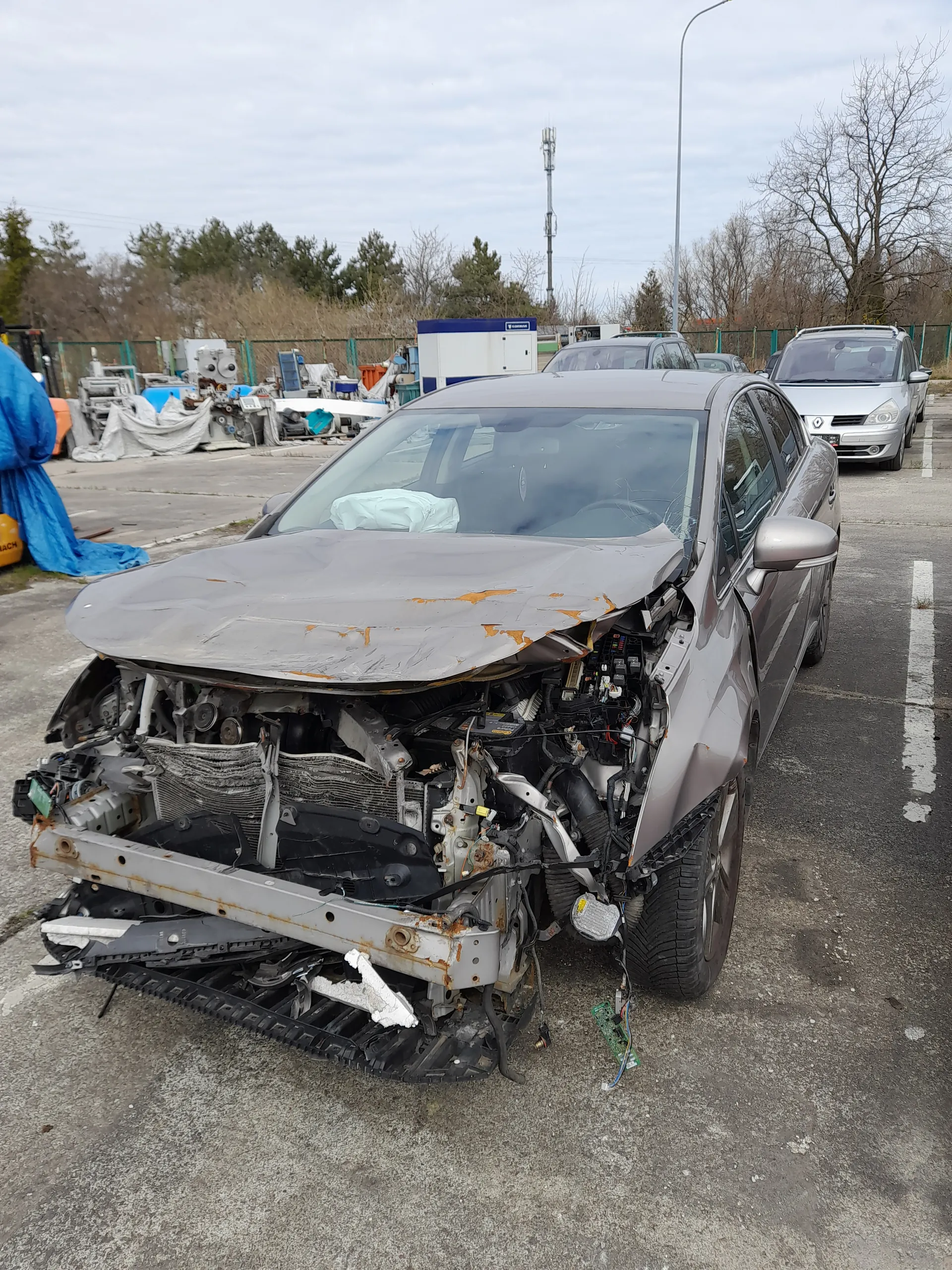 Zniszczony srebrny samochód osobowy z otwartą maską, ujawniającą uszkodzone elementy silnika i przedniej części. Widoczne są pęknięcia, rdza oraz rozrzucone części mechaniczne. W tle stoją inne pojazdy, w tym biały samochód, oraz teren z drzewami, ogrodzeniem i narzędziami przykrytymi niebieskimi i białymi płachtami. Niebo jest zachmurzone, a asfaltowa powierzchnia ma białe linie drogowe.