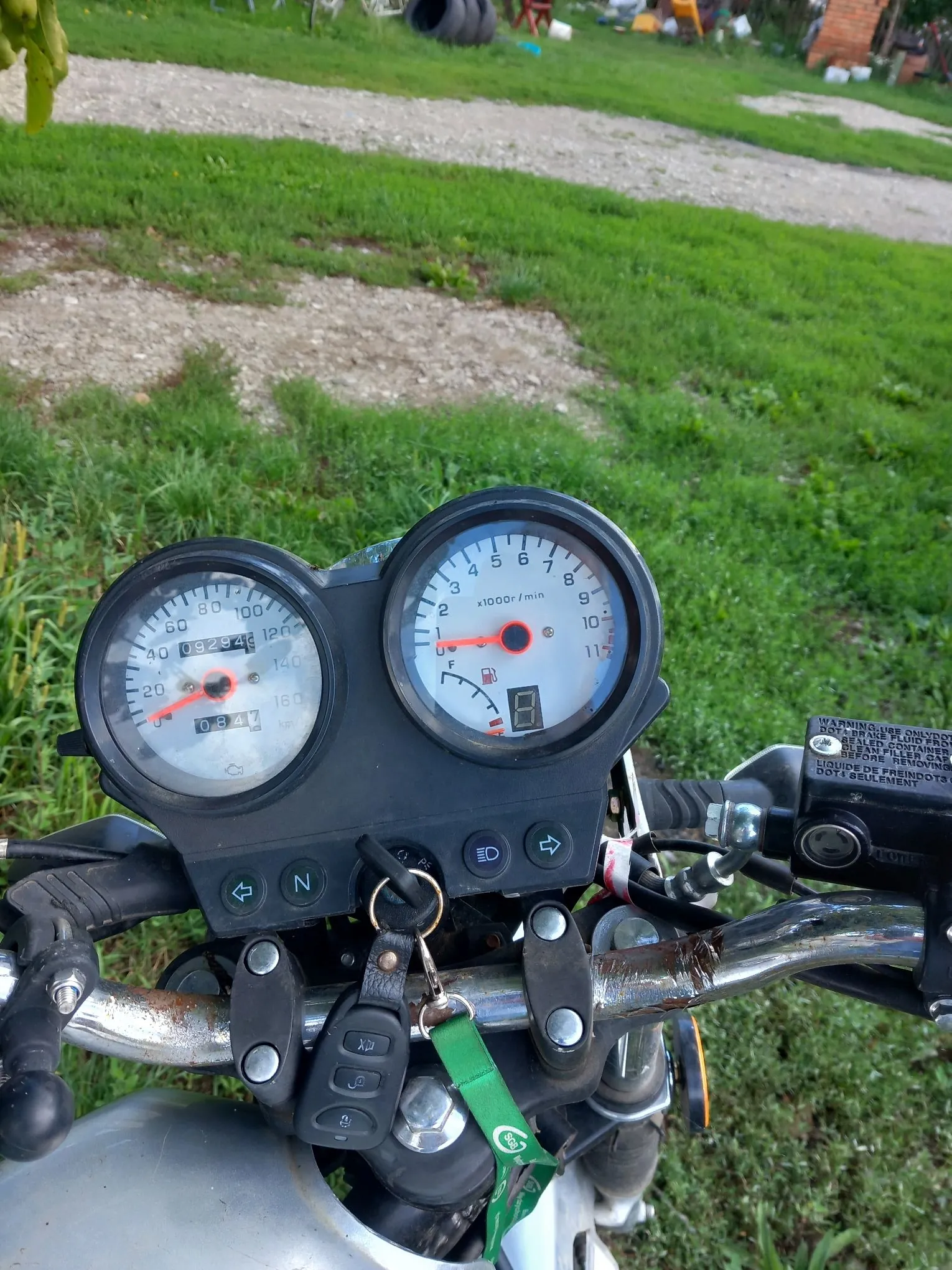 Widok z perspektywy kierowcy na kokpit motocykla. Widoczne są dwa okrągłe analogowe wskaźniki: po lewej prędkościomierz z cyfrowym licznikiem kilometrów (wartość 09294), po prawej obrotomierz z cyfrowym wyświetlaczem. Poniżej wskaźników znajduje się panel z kontrolkami i przyciskami, w tym przyciski kierunkowskazów i neutralnego biegu (N). Do stacyjki włożone są kluczyki z zieloną zawieszką. Kierownica jest metalowa, z widocznymi śladami rdzy. Tłem jest zielona trawa i żwirowa droga.