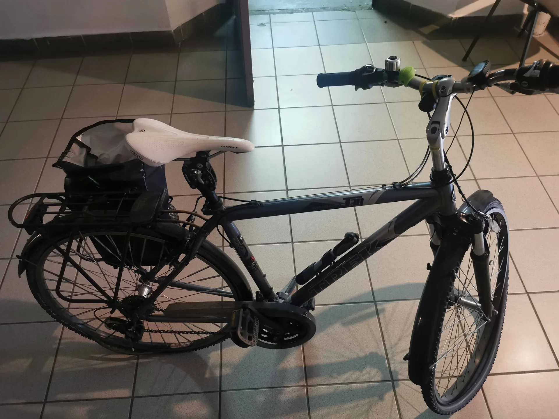 Czarny rower trekkingowy z szarą ramą, wyposażony w tylny bagażnik z zamontowaną białą torbą, błotniki oraz przedni amortyzator. Rower stoi bokiem na kafelkowej podłodze w pomieszczeniu.