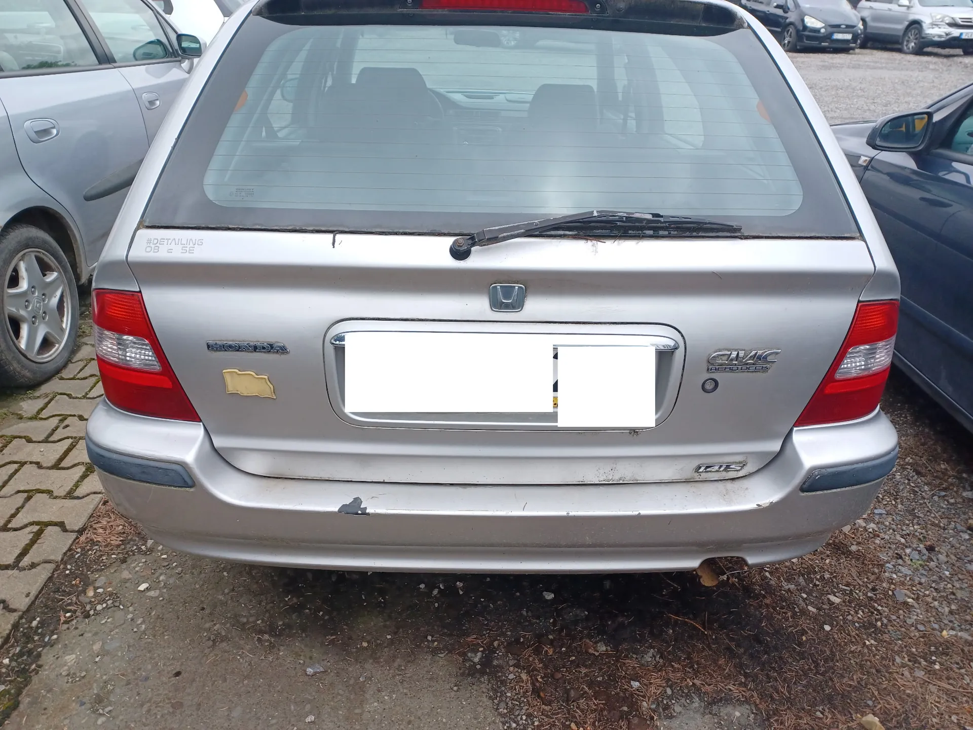 Tylny widok srebrnego samochodu marki Honda Civic Aerodeck. Na klapie bagażnika widoczne są napisy 'HONDA' po lewej stronie oraz 'CIVIC AERODECK' po prawej. Tablica rejestracyjna jest zakryta białym polem. Samochód posiada widoczne uszkodzenia w postaci wgnieceń i zarysowań na tylnym zderzaku. W tle po lewej stronie widoczny jest fragment innego srebrnego auta, a po prawej ciemnoniebieskiego samochodu.