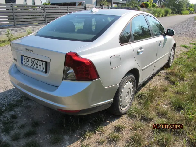 Srebrny samochód osobowy marki Volvo widziany od tyłu i z boku, zaparkowany na nieutwardzonym terenie obok trawy. Pojazd posiada polską tablicę rejestracyjną o numerze KR 896HN. W tle widoczny jest szary płot oraz domy.