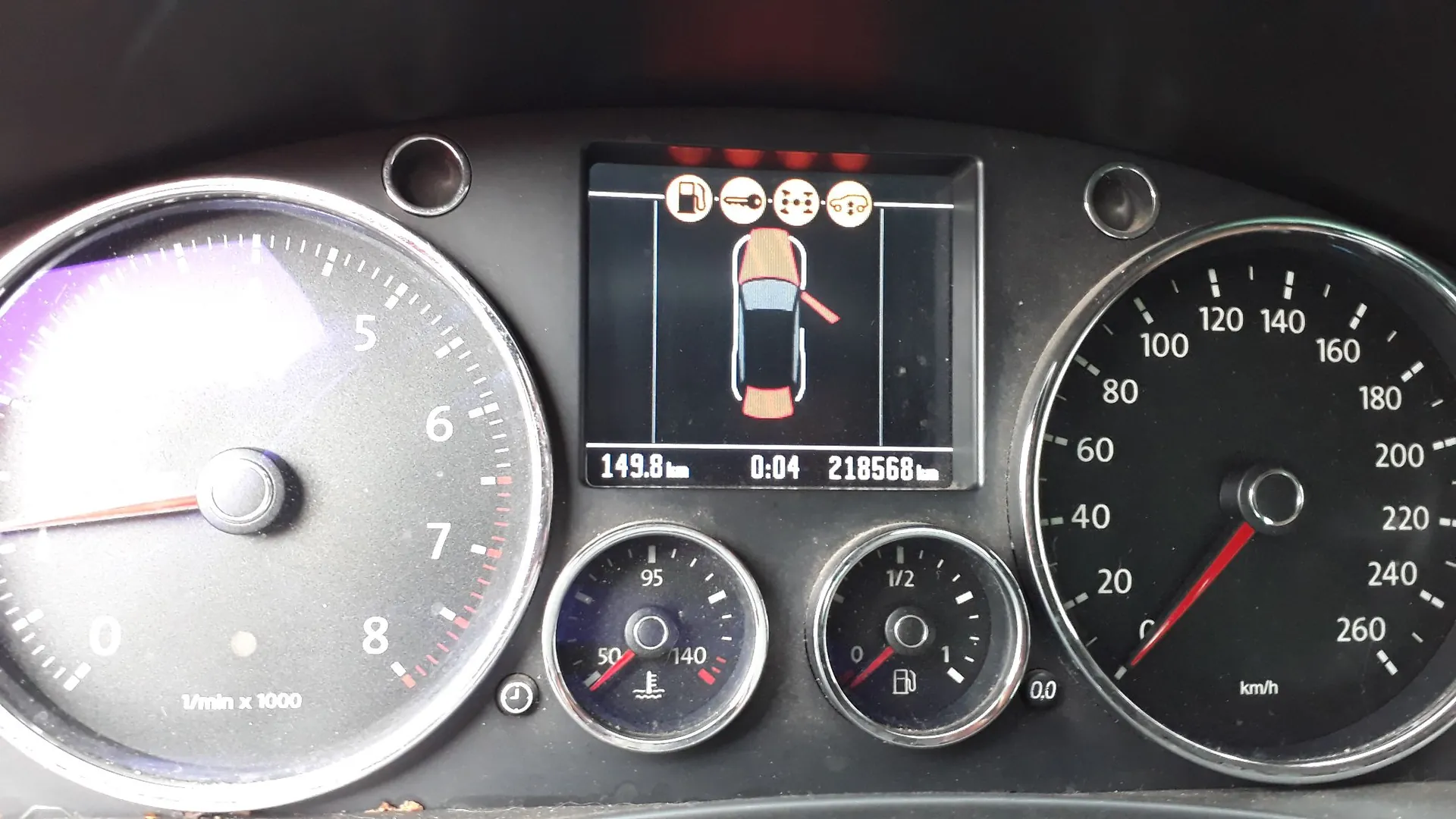 Panel instrumentów samochodu. Po lewej znajduje się obrotomierz z zaznaczonymi wartościami od 0 do 8 (1/min x 1000). Po prawej prędkościomierz z zakresem do 260 km/h. W centrum znajduje się ekran cyfrowy wyświetlający schemat samochodu z otwartymi drzwiami pasażera oraz wskaźnikami ostrzegawczymi (olej, silnik, oświetlenie). Na dole ekranu widoczne są wartości: 149.8 km, 0:04 oraz przebieg 218568 km. Poniżej ekranu znajdują się dwa mniejsze wskaźniki: temperatura płynu chłodniczego (zakres 50-140) oraz poziom paliwa (zakres 0-1/2).