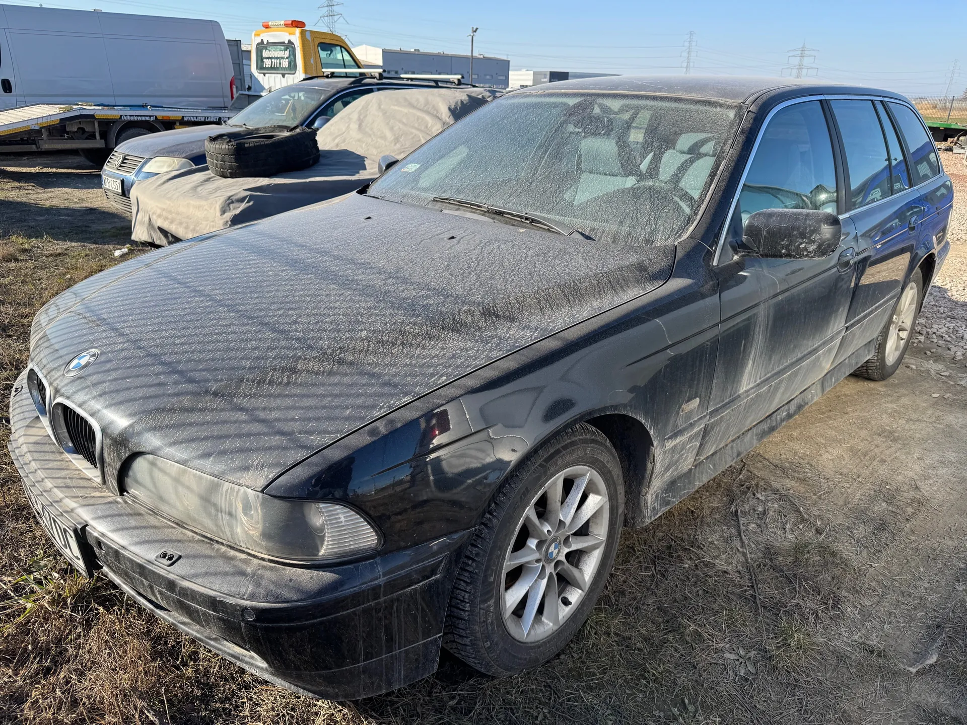 Czarny samochód osobowy marki BMW kombi pokryty kurzem, zaparkowany na suchym trawniku i żwirze. W tle widoczne są inne pojazdy: zakryty płachtą samochód z oponą na dachu, niebieski samochód, biały van oraz żółty wóz holowniczy z napisem "odholowanie.pl" i numerem telefonu "790 711 166". Na horyzoncie budynki przemysłowe i linie energetyczne. Niebo jest bezchmurne.