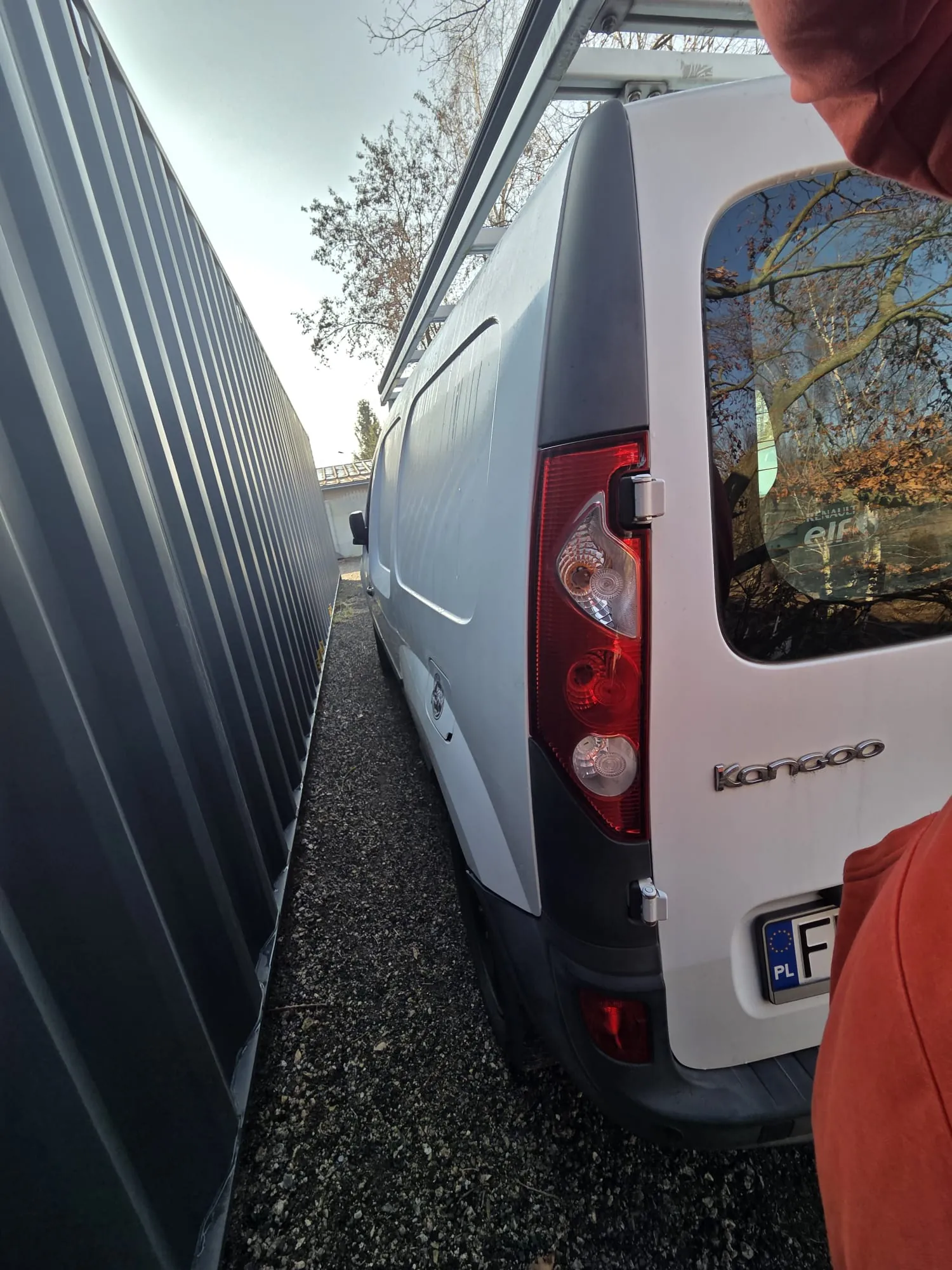 Biały samochód dostawczy Renault Kangoo zaparkowany obok szarego kontenera. Widoczne są tylna część pojazdu z czerwonymi światłami, napisem 'Kangoo' oraz tablicą rejestracyjną zaczynającą się na 'PL F'. Po prawej stronie fragment pomarańczowej kurtki osoby. Tło: drzewa bez liści, budynek i niebo. Podłoga z drobnej kostki.