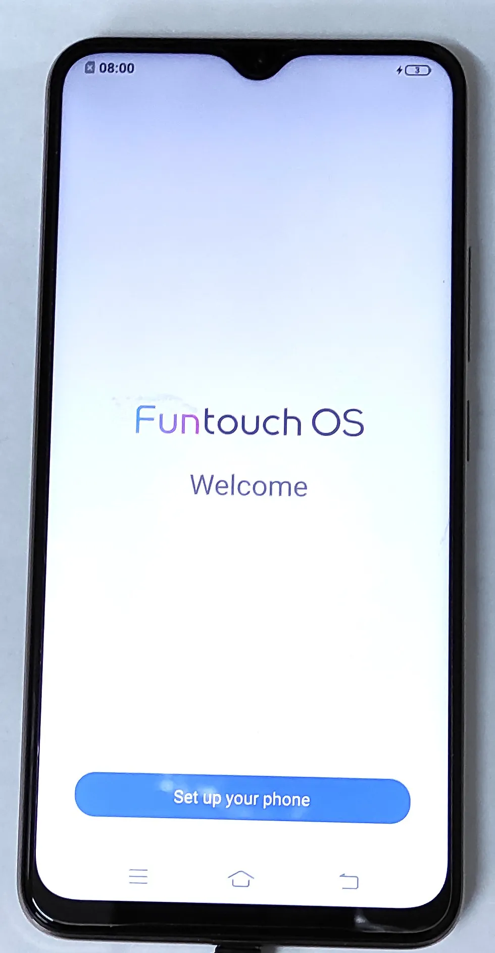 Smartfon z włączonym ekranem wyświetlającym stronę powitalną systemu operacyjnego. Na białym tle z delikatnym błękitnym gradientem widnieje napis „Funtouch OS” oraz poniżej słowo „Welcome”. Na dole ekranu znajduje się niebieski przycisk z napisem „Set up your phone”. W górnym pasku widoczna jest godzina 08:00 oraz ikona baterii z poziomem naładowania 3%. Na samym dole znajdują się systemowe przyciski nawigacyjne.