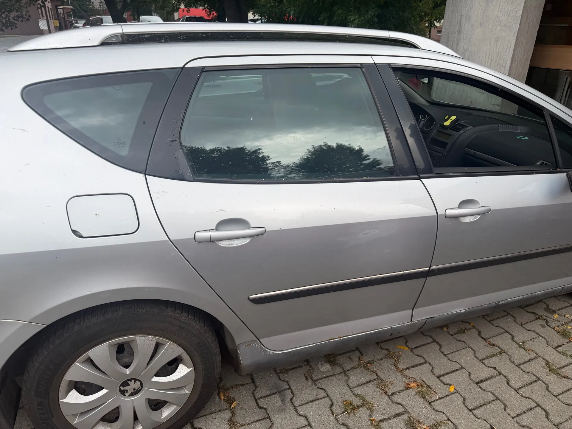 Srebrny samochód typu kombi zaparkowany na kostce brukowej. Widoczny jest bok pojazdu z dwiema drzwiami, kołem z plastikową osłoną oraz klapką wlewu paliwa. Na drzwiach widoczne są drobne zarysowania i wgniecenia.