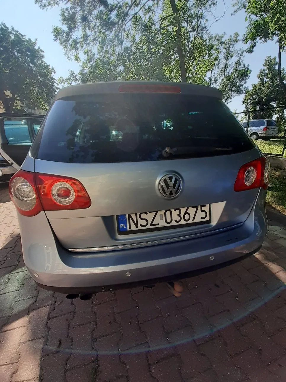 Srebrny samochód marki Volkswagen widziany od tyłu, zaparkowany na kostce brukowej. Na samochodzie znajduje się polska tablica rejestracyjna o numerze NSZ 03675. W tle widoczne są zielone drzewa i fragment nieba.
