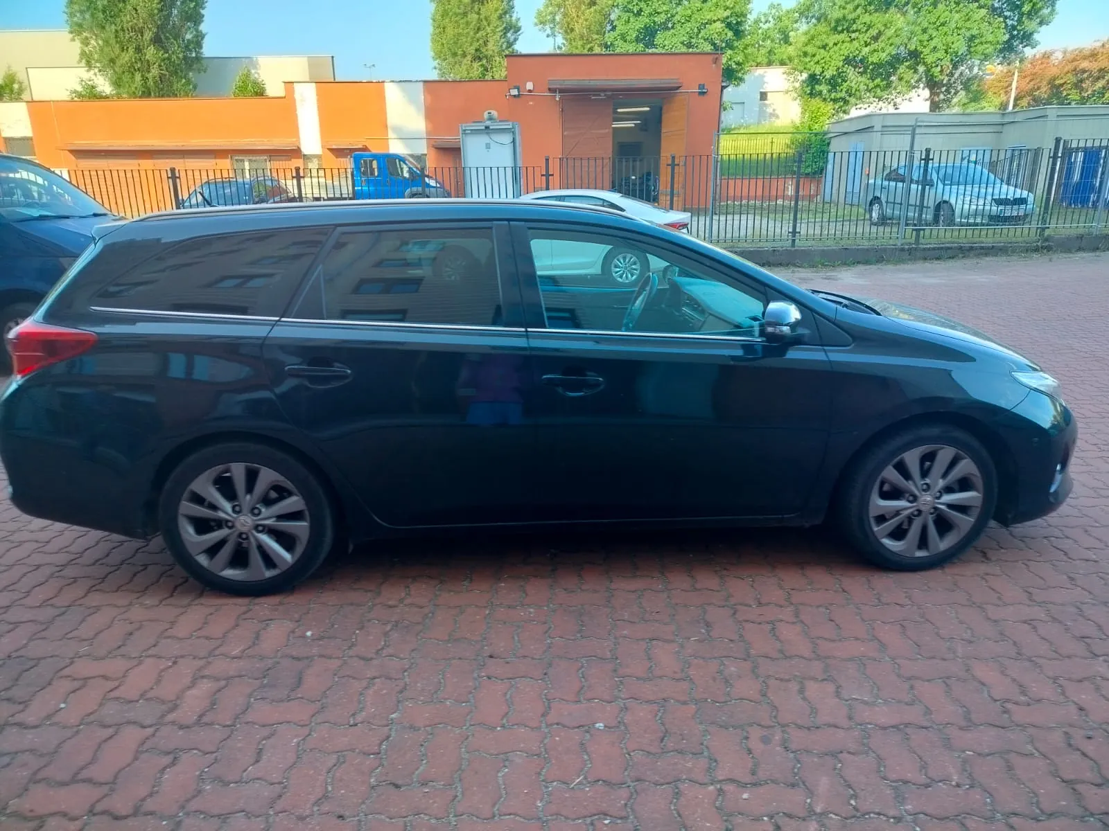 Czarny samochód typu kombi zaparkowany na czerwonej brukowanej powierzchni. W tle widoczne są pomarańczowe i beżowe budynki, metalowe ogrodzenie, inne zaparkowane pojazdy (w tym niebieski samochód dostawczy i srebrny osobowy) oraz drzewa. Niebo jest jasne, a oświetlenie wskazuje na słoneczny dzień.