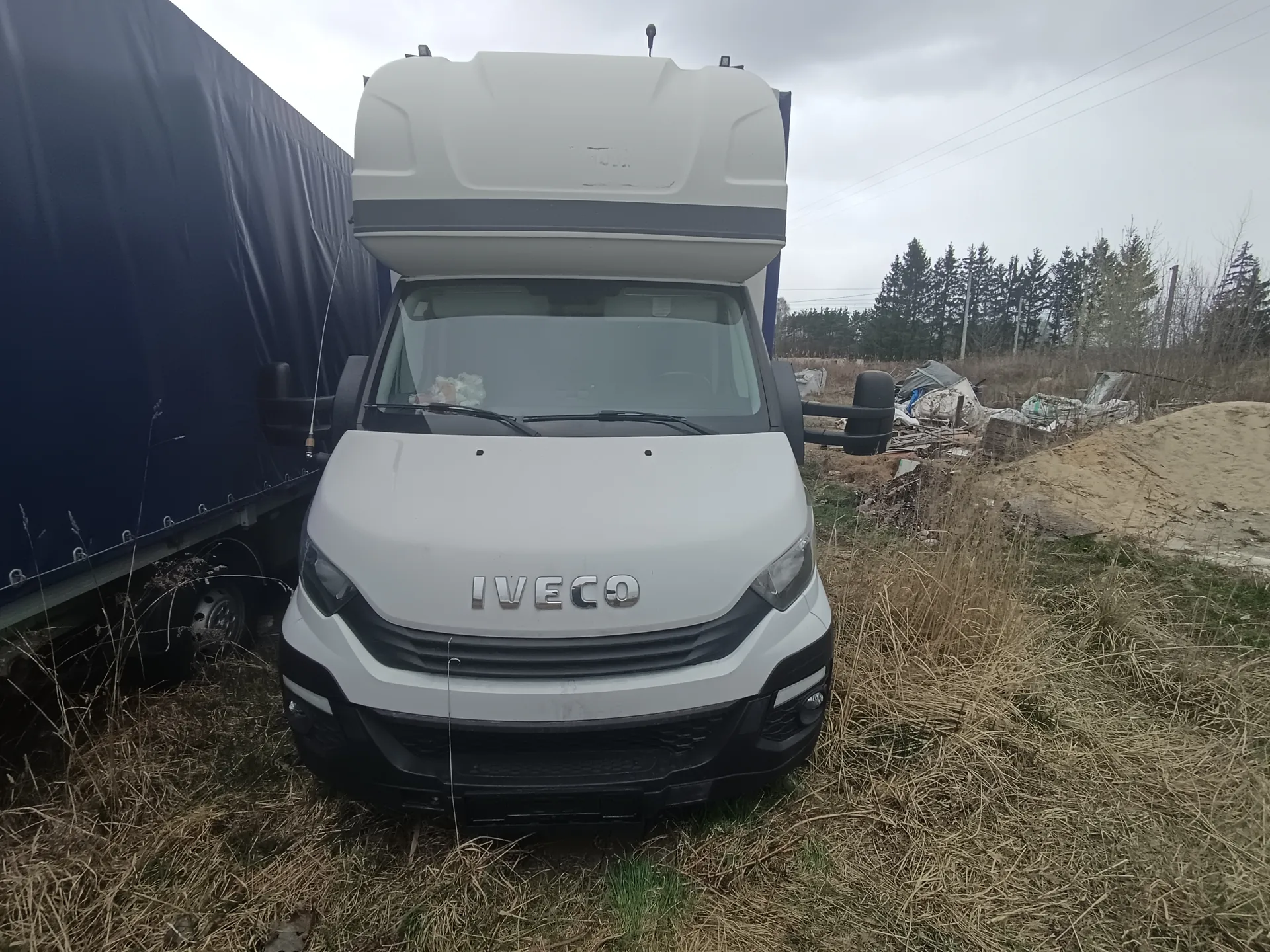 Biały samochód dostawczy marki IVECO z widocznym napisem na masce, stojący na trawie. Po lewej stronie obrazu znajduje się niebieski ciężarówka z przyczepą. W tle widoczne są suche trawy, sterta śmieci, kopiec piasku, drzewa iglaste oraz pochmurne niebo. Na przedniej szybie samochodu IVECO leży biały przedmiot.