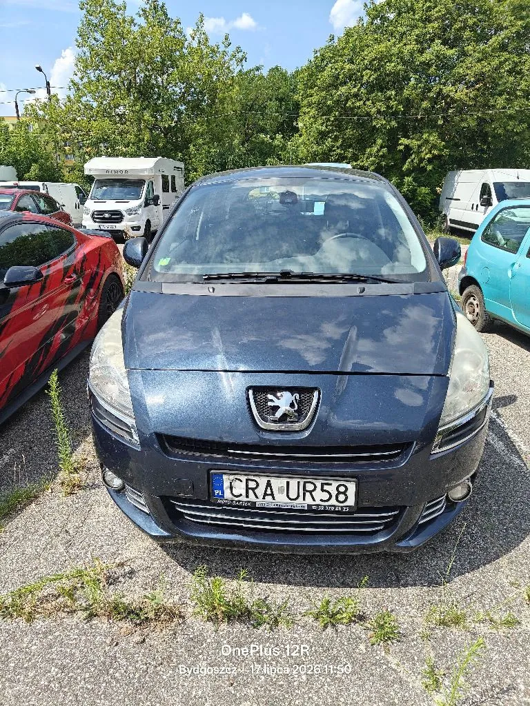 Zdjęcie przodu ciemnoniebieskiego samochodu marki Peugeot zaparkowanego na żwirowym placu. Pojazd ma tablicę rejestracyjną o numerze CRA UR58. W tle widoczne są inne samochody, w tym czerwony sportowy samochód po lewej stronie, biały kamper oraz jasnoniebieski samochód po prawej stronie. W tle znajdują się zielone drzewa i jasne niebo. Na dole zdjęcia widnieje biały napis: OnePlus 12R Bydgoszcz, 17 lipca 2024, 11:50.