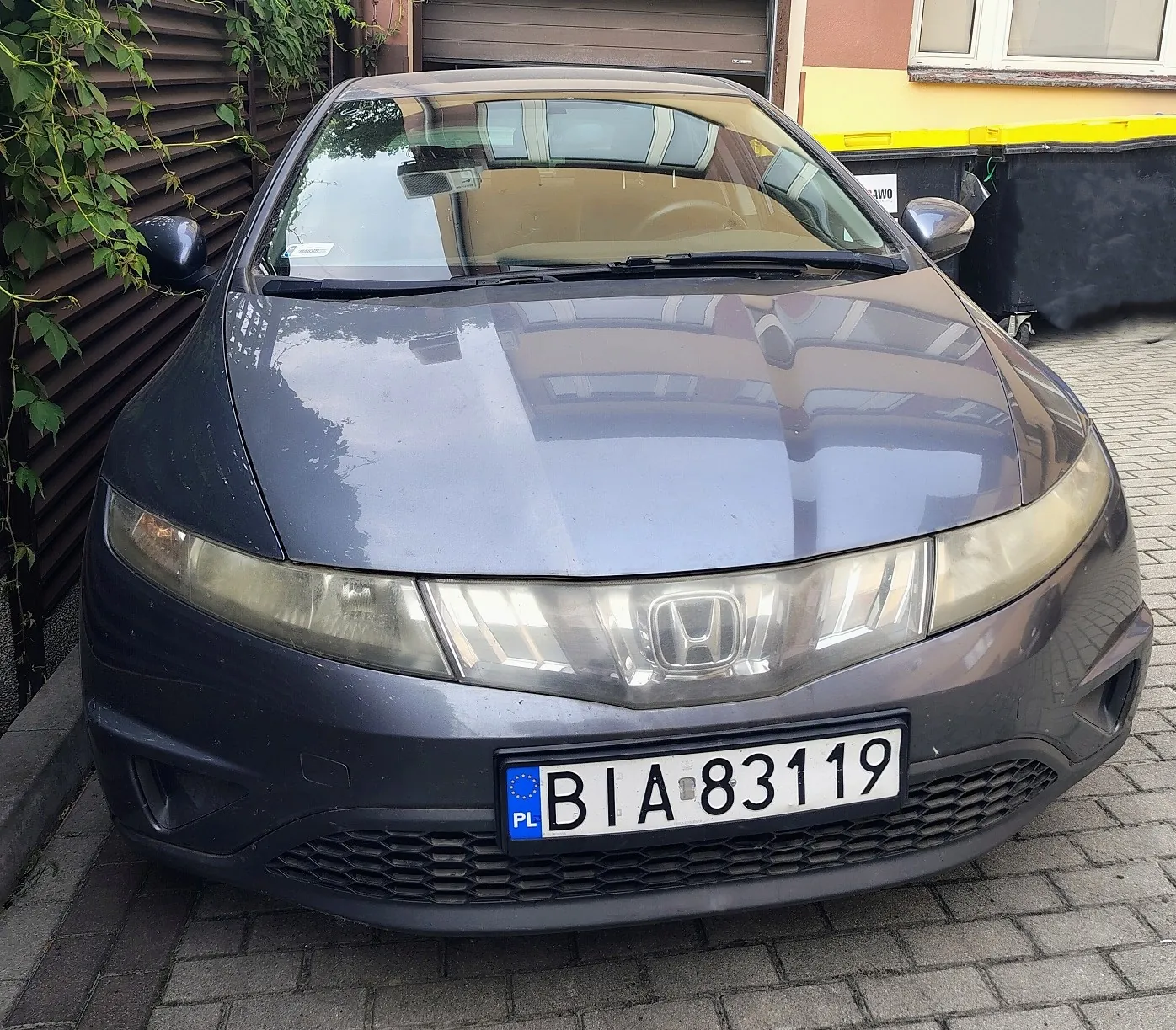 Szary samochód marki Honda zaparkowany na kostce brukowej. Widoczny jest przód pojazdu z tablicą rejestracyjną o numerze BIA 83119. W tle znajduje się budynek z brązową bramą garażową oraz zielone pnącza na ścianie po lewej stronie.