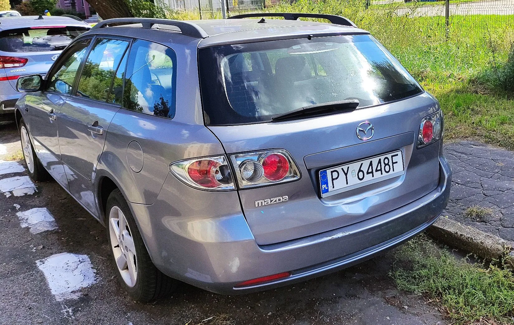 Srebrnoszary samochód marki Mazda w nadwoziu kombi, zaparkowany tyłem do obiektywu na utwardzonym podłożu. Pojazd posiada polską tablicę rejestracyjną o numerze PY 6448F. Na tylnej klapie widoczne jest logo Mazdy oraz napis 'mazda'. Samochód ma zamontowane relingi dachowe.