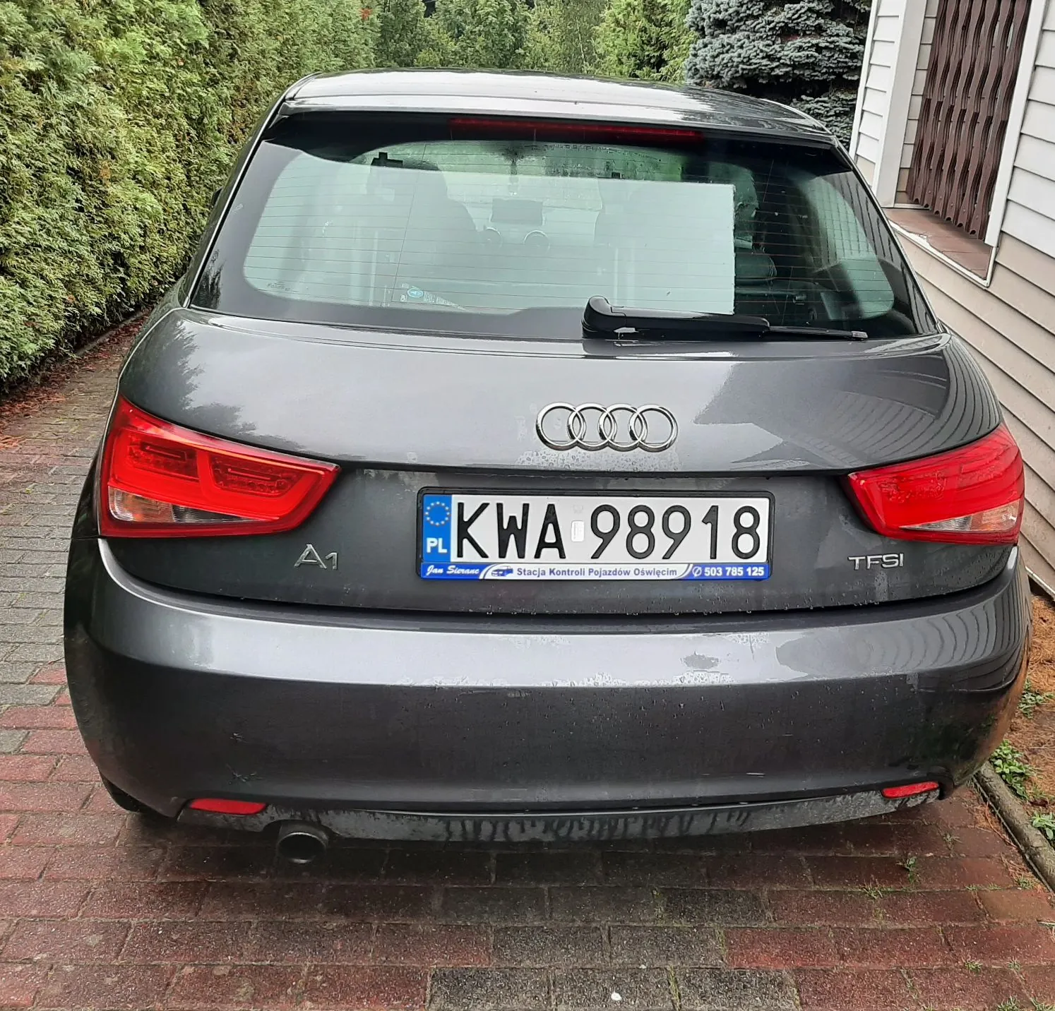 Tylny widok szarego samochodu marki Audi A1. Na klapie bagażnika znajduje się logo Audi, oznaczenie modelu A1 oraz napis TFSI. Samochód ma polską tablicę rejestracyjną o numerze KWA 98918. Poniżej tablicy widnieje napis: 'Jan Suraś Stacja Kontroli Pojazdów Oświęcim 503 785 125'. Auto stoi na kostce brukowej obok zielonego żywopłotu i fragmentu budynku.