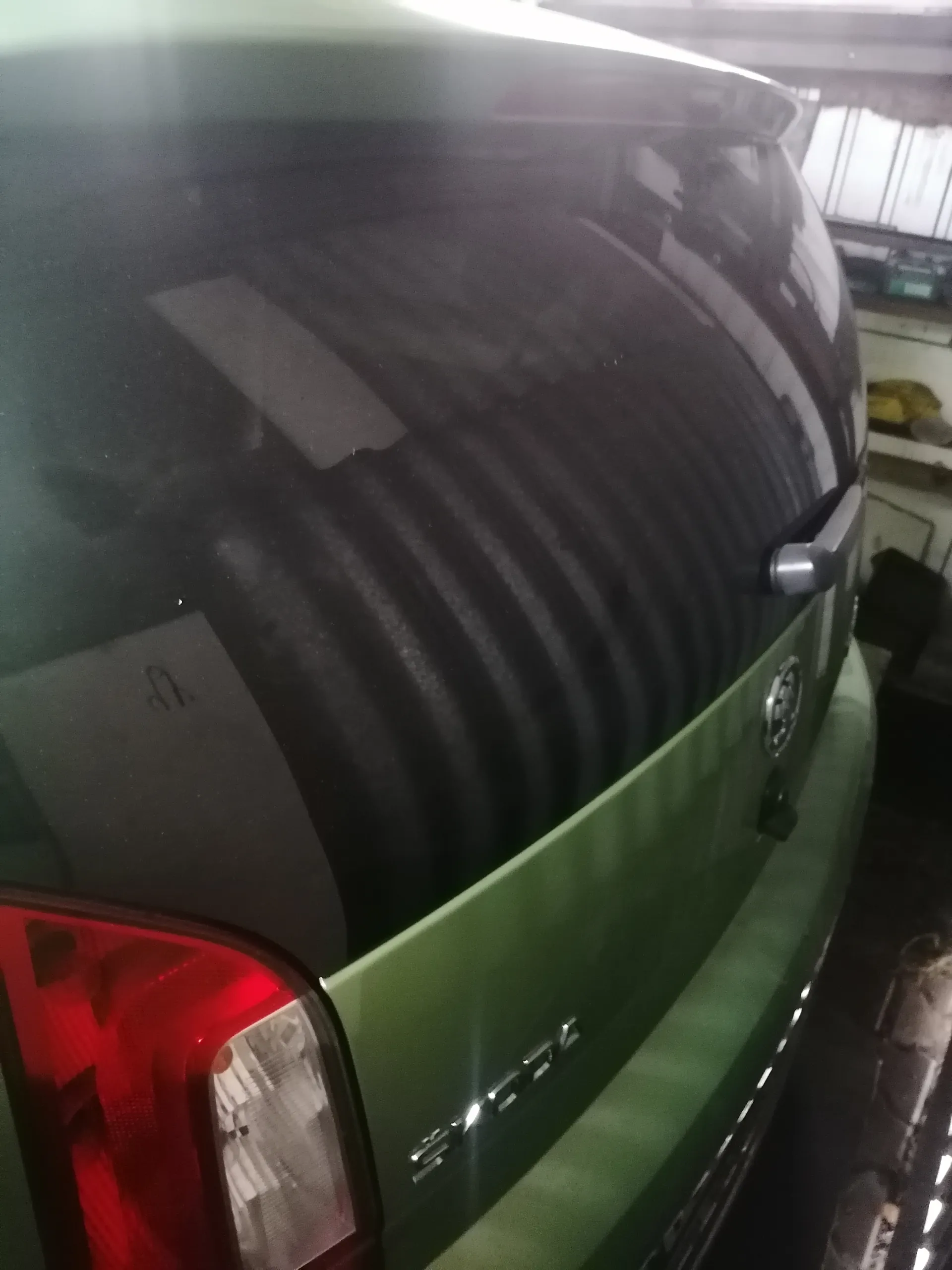 Zbliżenie na tył zielonego samochodu marki Suzuki. Widoczny czerwony reflektor, emblemat Suzuki na bagażniku oraz fragmenty wnętrza garażu w tle, w tym półki z narzędziami i przedmiotami. Na szybie tylnej znajduje się nieczytelny napis.