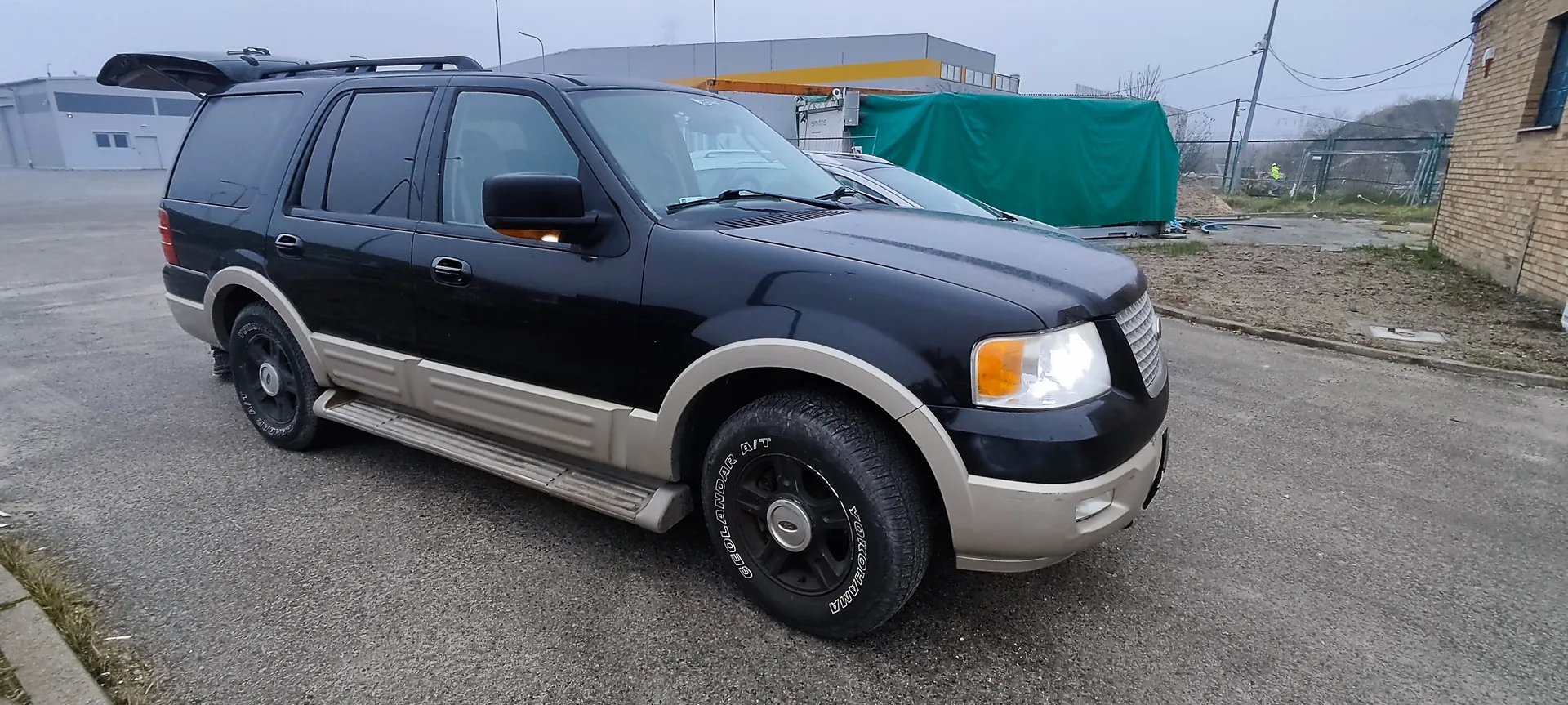 Czarny SUV z beżowymi elementami nadwozia zaparkowany na asfaltowej powierzchni. Tylna klapa pojazdu jest otwarta, widoczny dachowy bagażnik. Opony z napisem GEOLANDAR A/T YOKOHAMA. W tle budynek przemysłowy z żółtą listwą, zielona płachta, ceglany budynek oraz ogrodzenie. Niebo jest pochmurne.