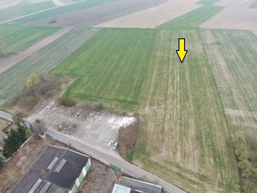 Zdjęcie lotnicze przedstawiające obszar rolniczy z polami o różnych odcieniach zieleni i brązu. W dolnej części obrazu widoczny jest betonowy plac z zaparkowanymi samochodami oraz fragmenty budynków przemysłowych. Na jednym z zielonych pól znajduje się żółta strzałka skierowana w dół.